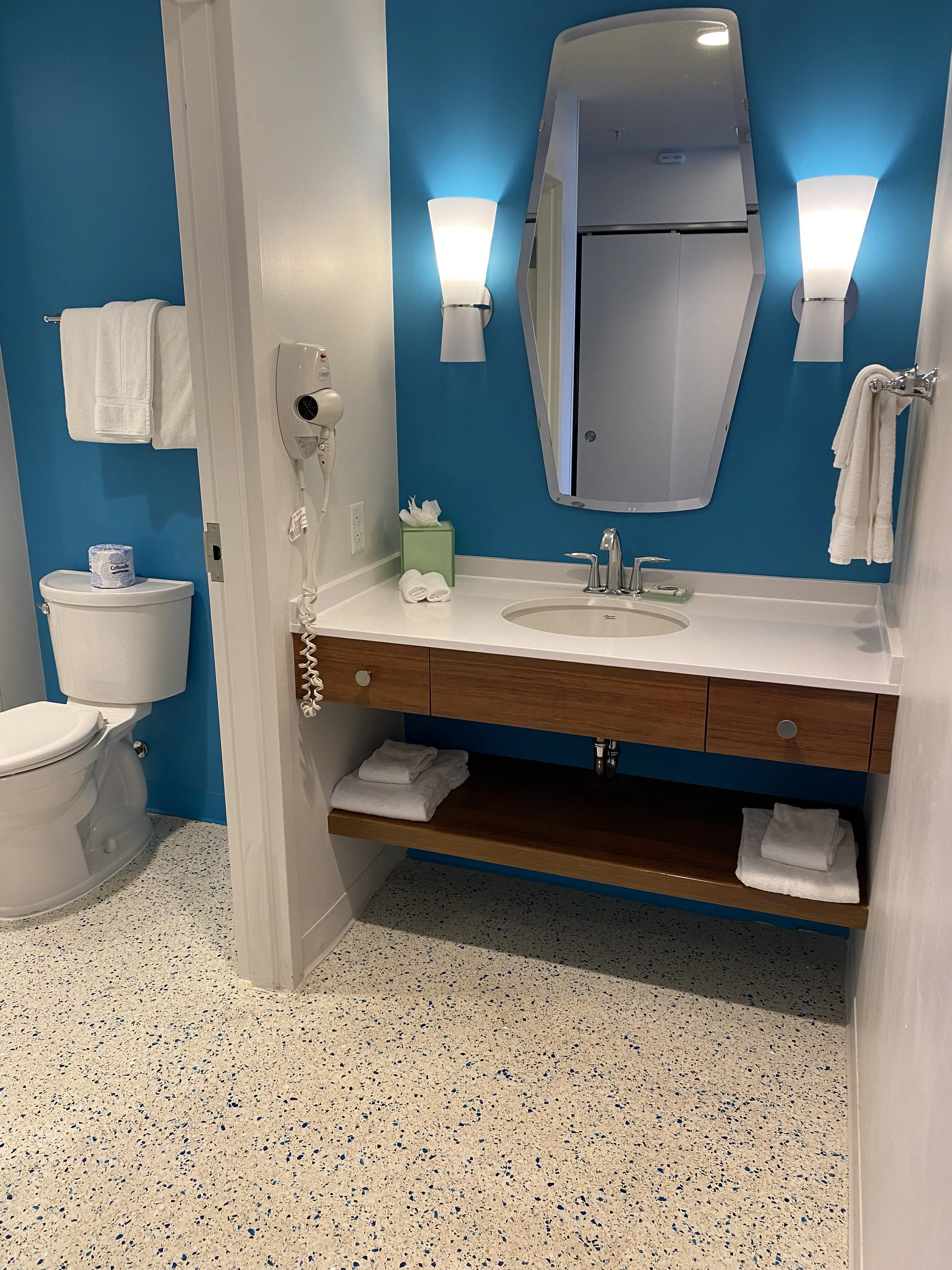 Bathroom with blue accent wall, white towels, a toilet, wall-mounted hairdryer, and a white sink with a wooden cabinet and mirror.