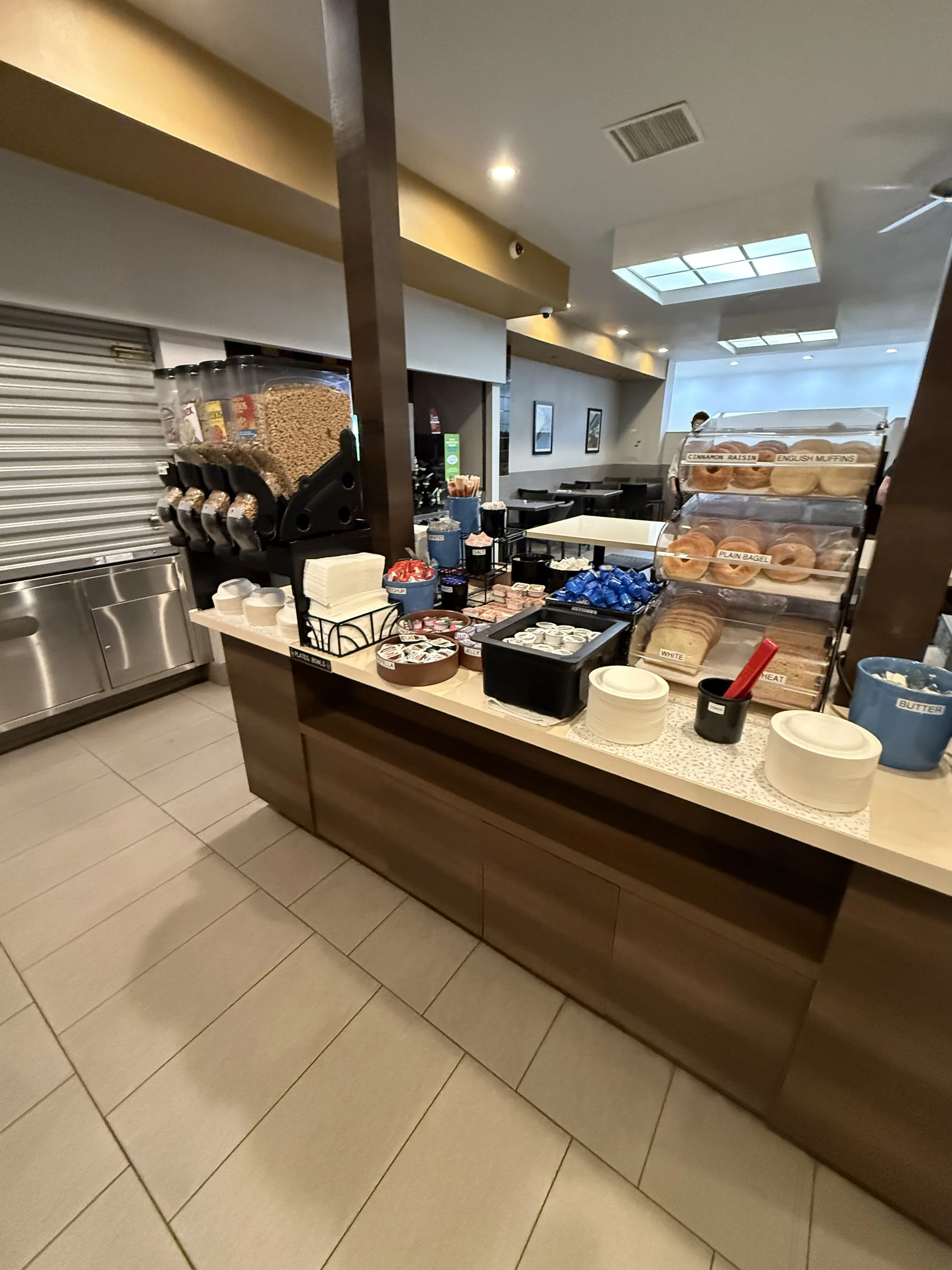 Breakfast buffet with cereal dispensers, bagels, muffins, butter, and various condiments on a counter in a dining area.