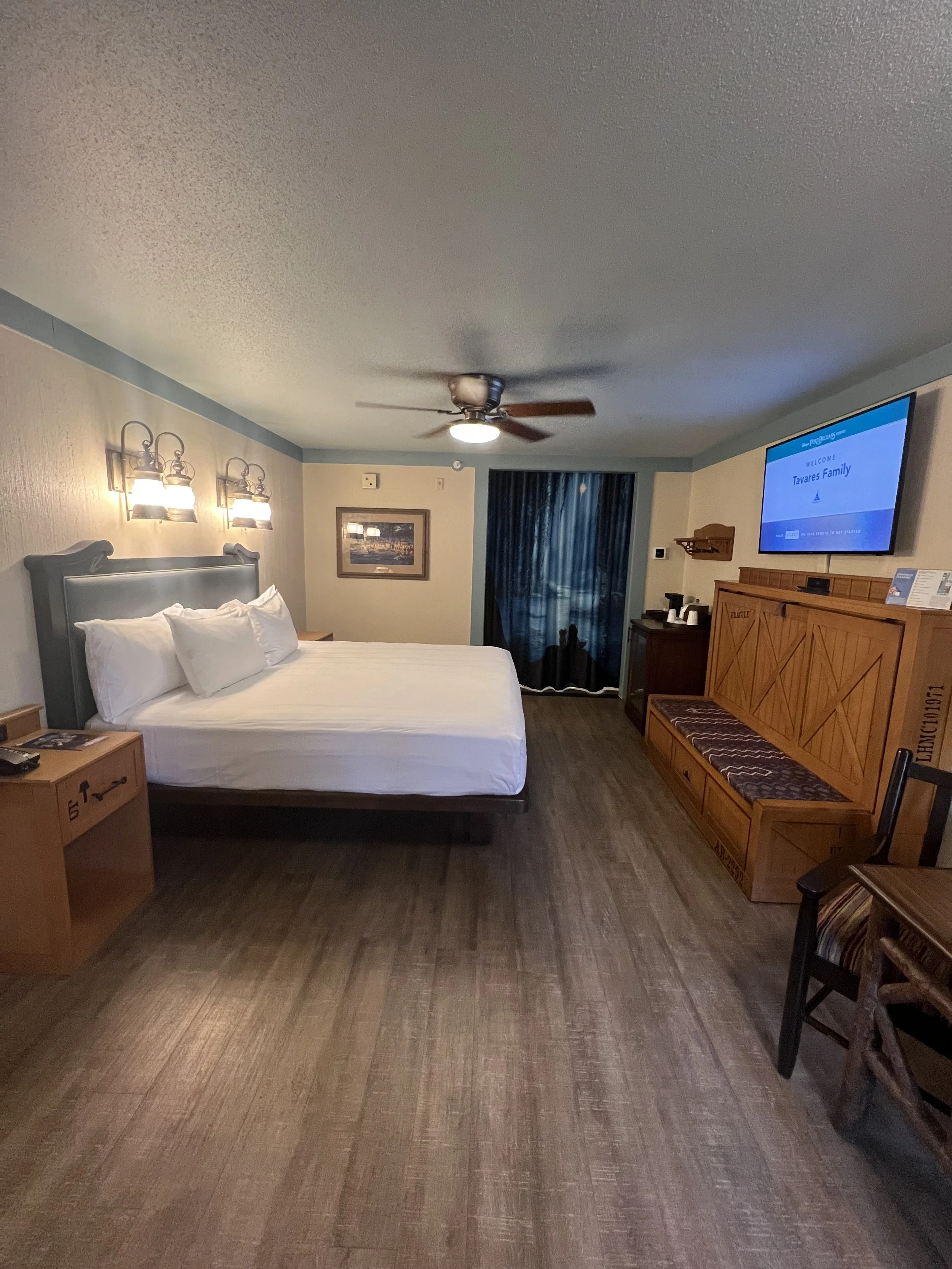 A hotel room with a large bed, wooden furniture, a ceiling fan, a wall-mounted TV displaying a welcome message, a window with dark curtains, a chair, and a small table with a telephone and other items.