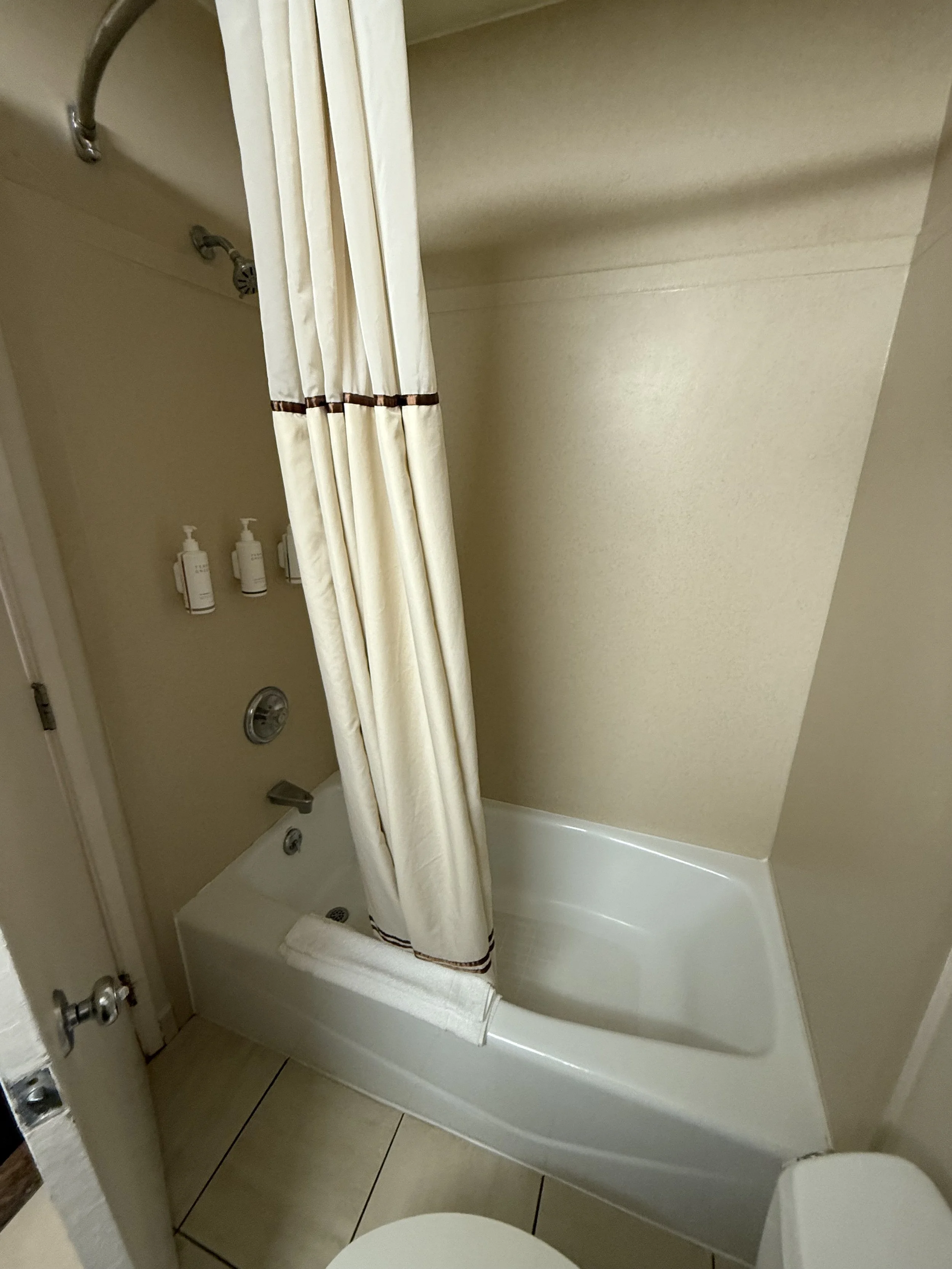 A small bathtub with a shower curtain, soap dispensers, and a showerhead in a beige bathroom.
