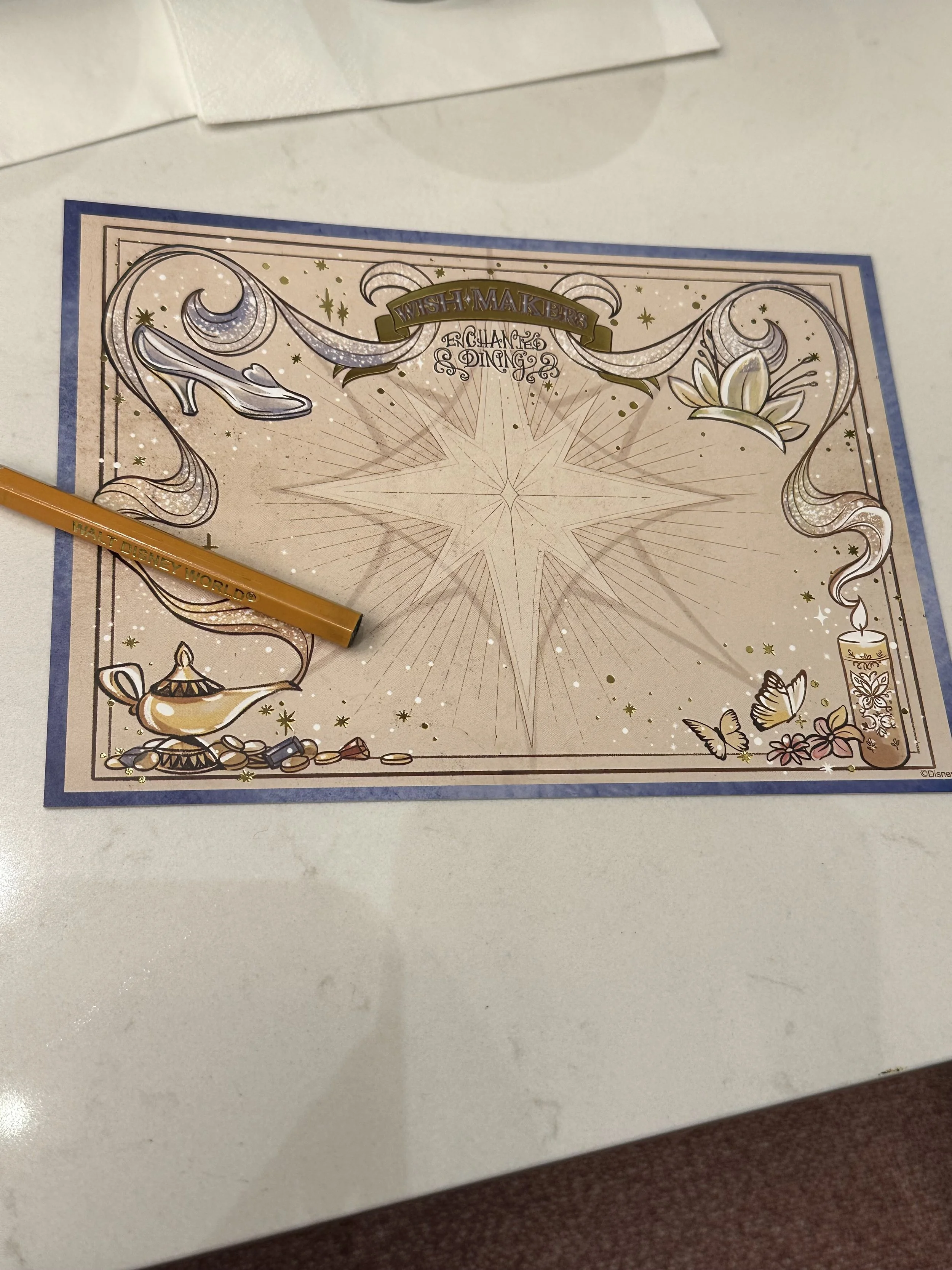 A Disney-themed wish-making activity sheet on a table with a gold-colored pencil labeled "WALT DISNEY WORLD." The sheet features a large star in the center with decorative elements including a lamp, butterflies, flowers, a candle, and a ribbon with t