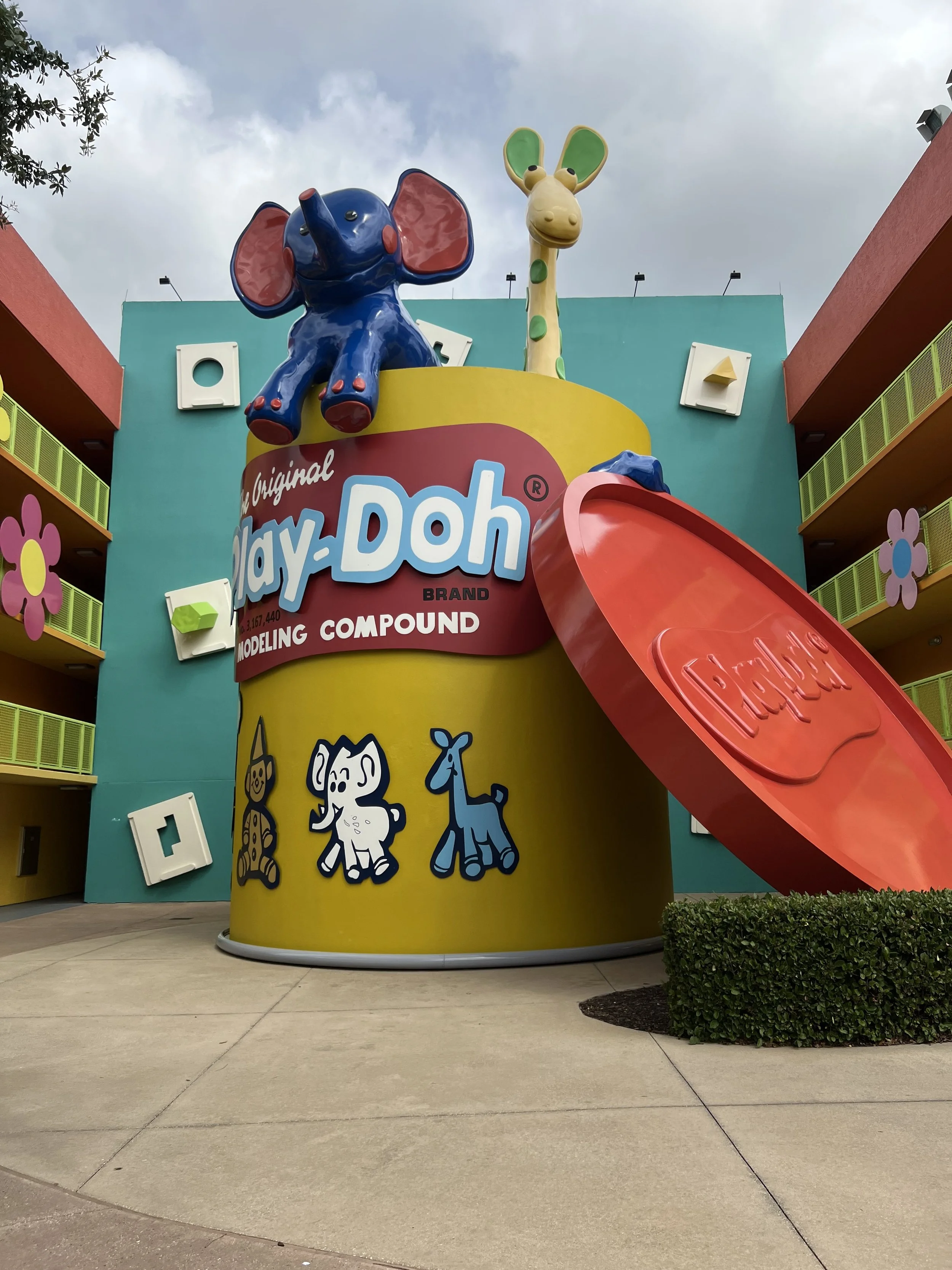 Large colorful Play-Doh model featuring an elephant and a giraffe on top, with a big red slide attached. The model is decorated with cartoon animal illustrations and is part of a themed building with bright pink flowers and green balconies.