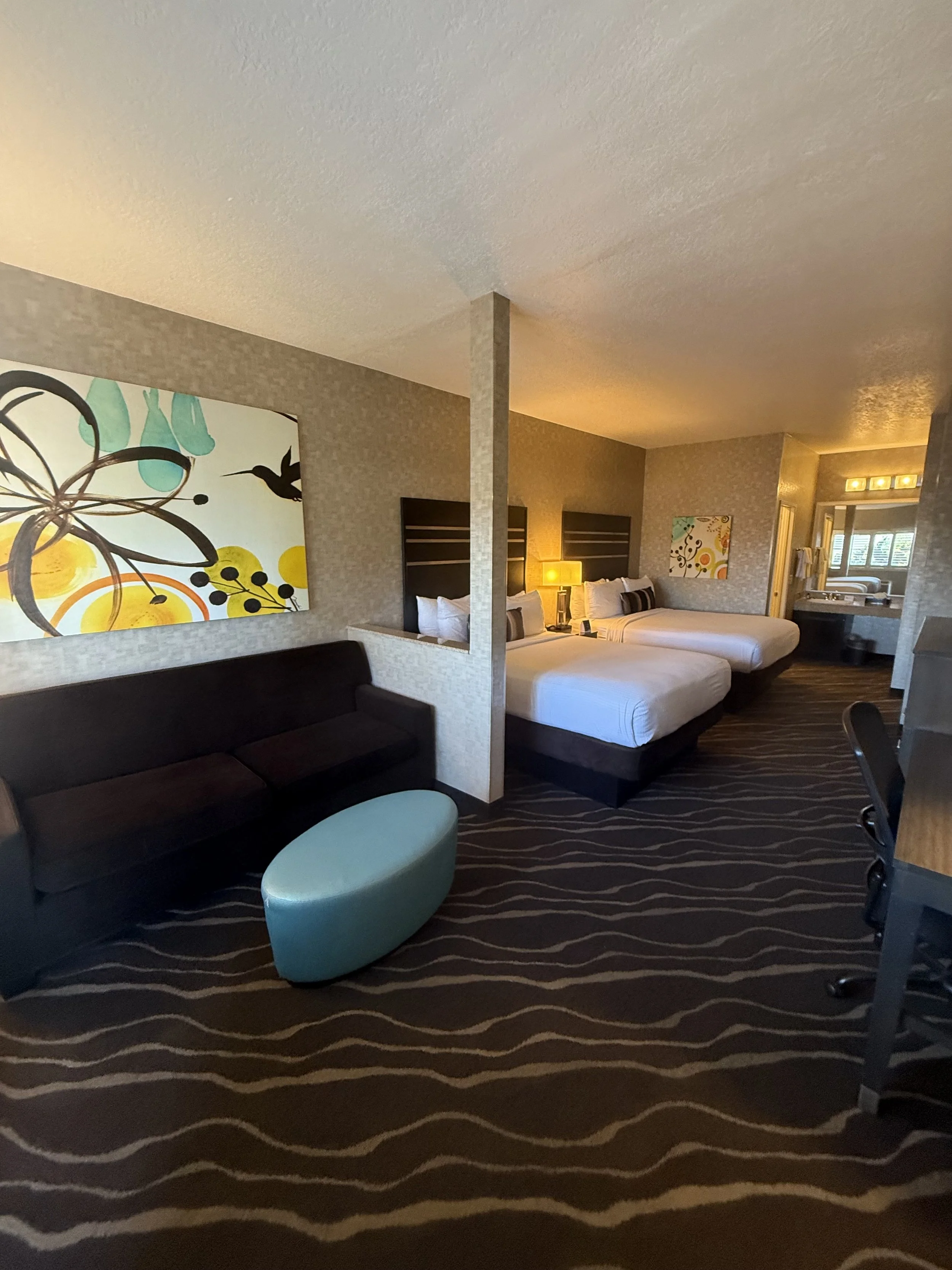 Hotel room with two beds, a dark sofa, a blue ottoman, and wall art. A lamp and a window in the background.