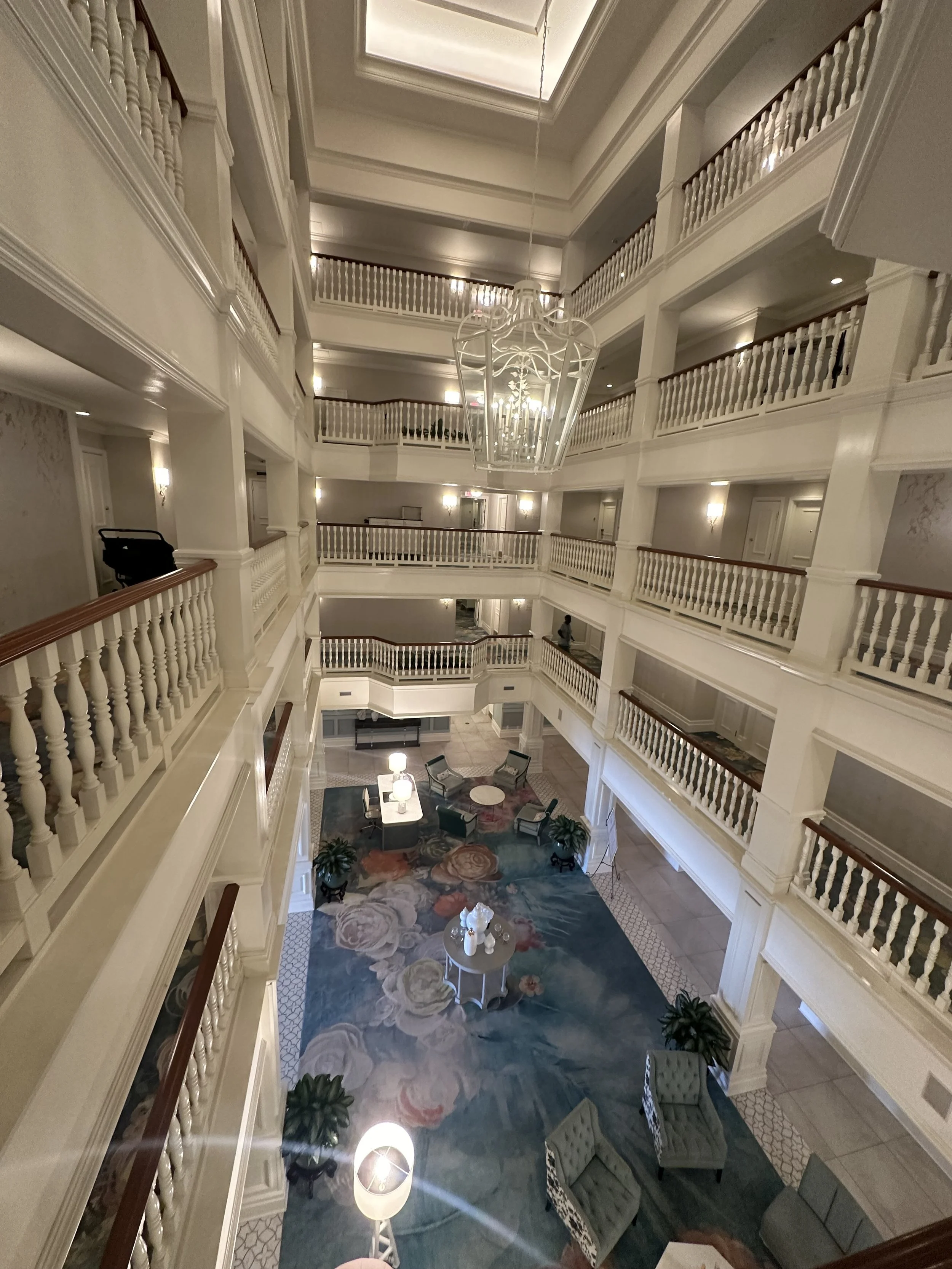 Multiple floors of a hotel interior with a high ceiling, chandelier, and a spacious lobby area with seating and a floral rug, viewed from a higher floor.