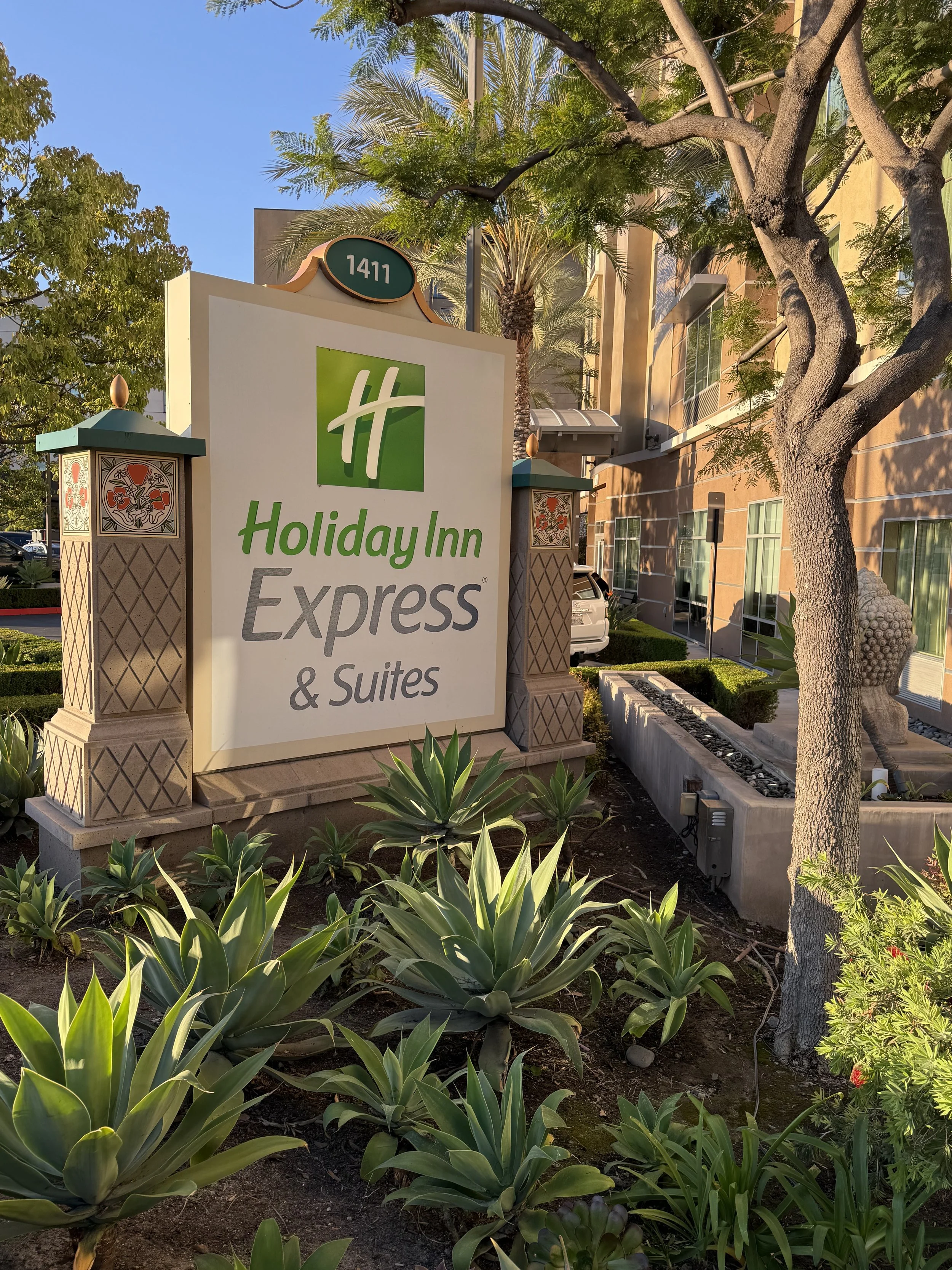 The image shows the entrance sign of a Holiday Inn Express & Suites hotel, with the address 1411. Surrounding the sign are desert plants, a tree, and some bushes. There are buildings and cars in the background, with clear skies overhead.