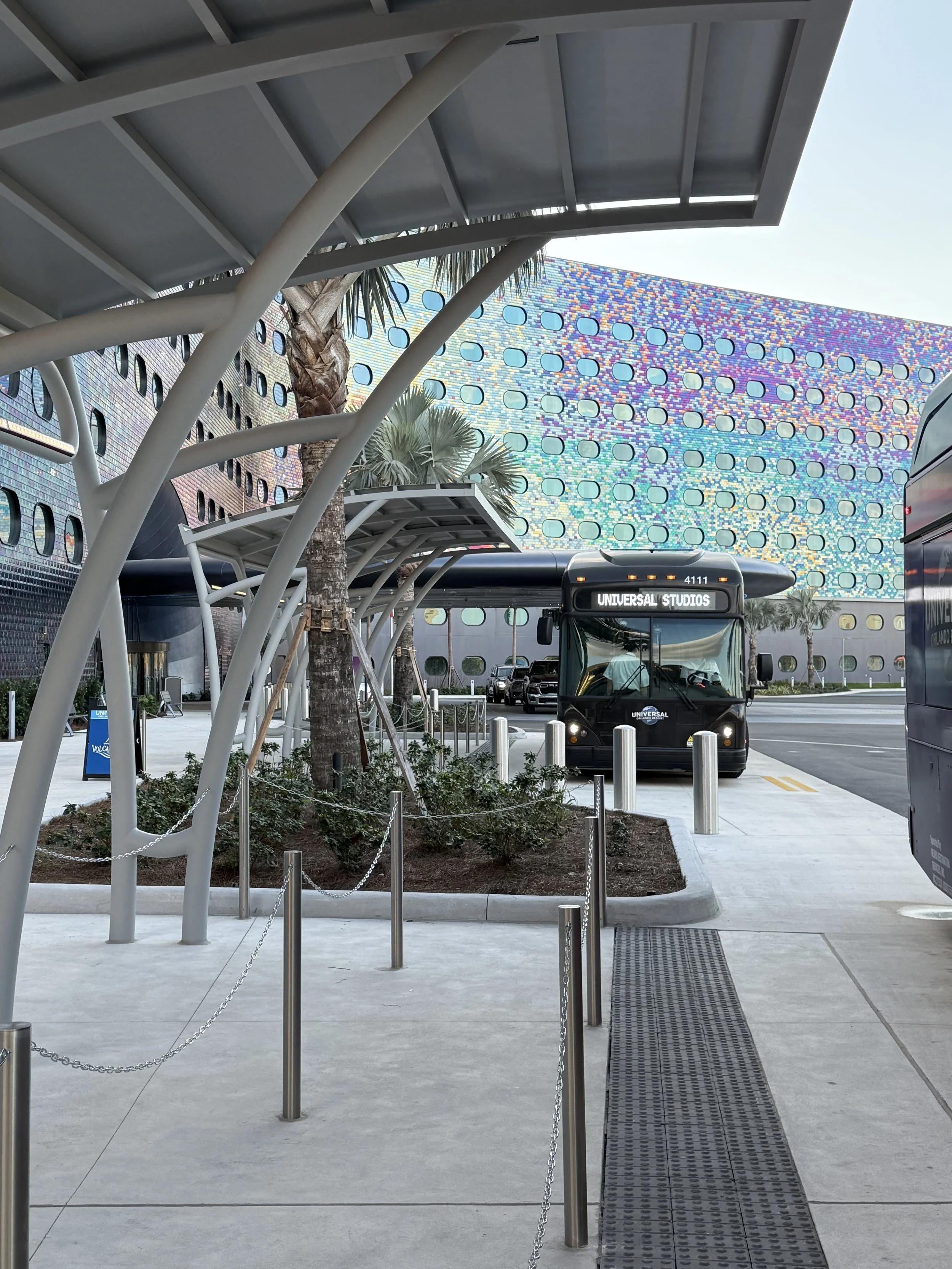 A black shuttle bus labeled 'Universal Studios' parked at a modern, colorful building with a tiled, iridescent exterior. There are palm trees, a small garden, and metal posts with chains guiding pedestrians in the foreground.