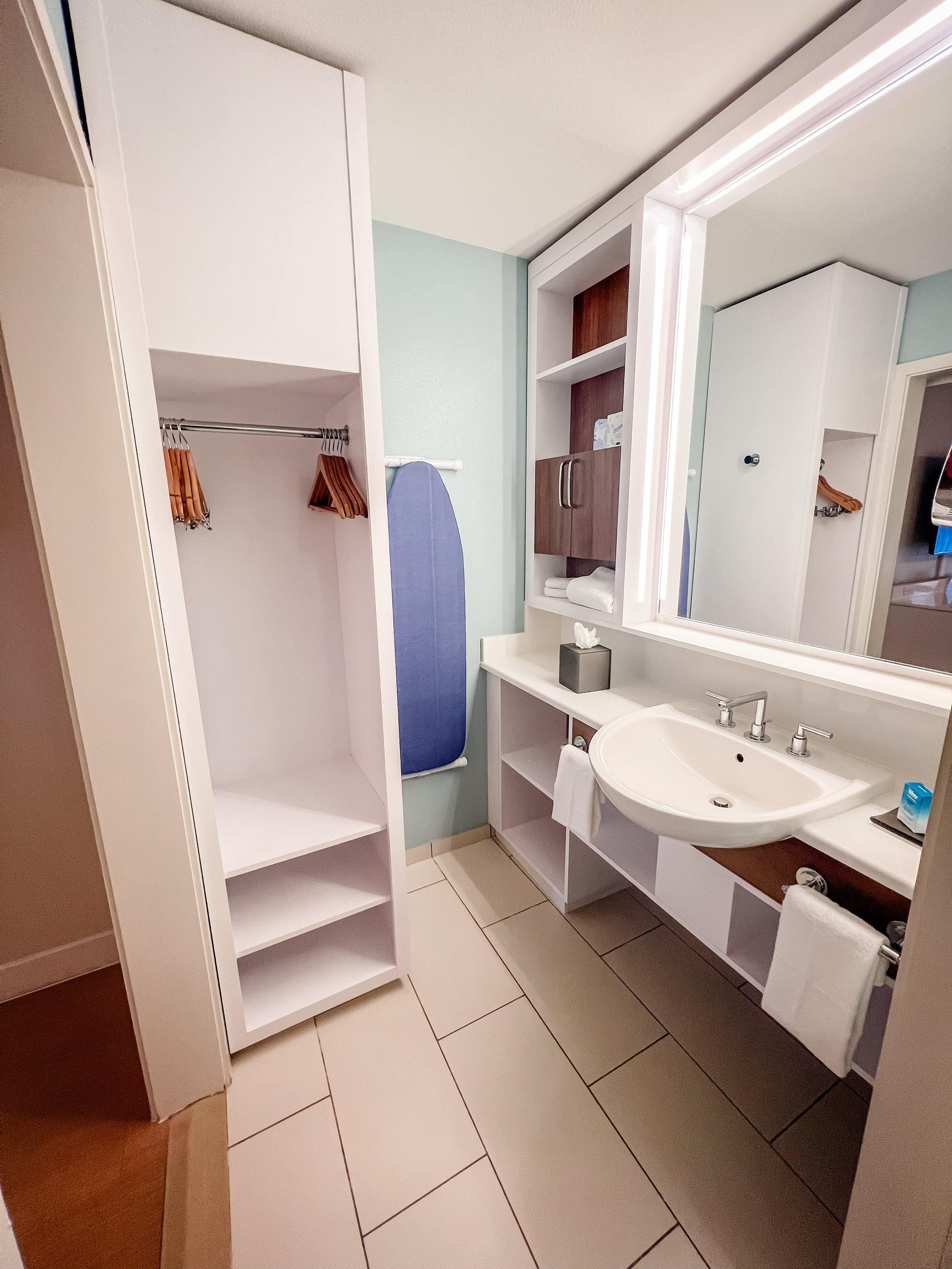 Bathroom with a white sink, mirror, shelves, closet, and an ironing board.