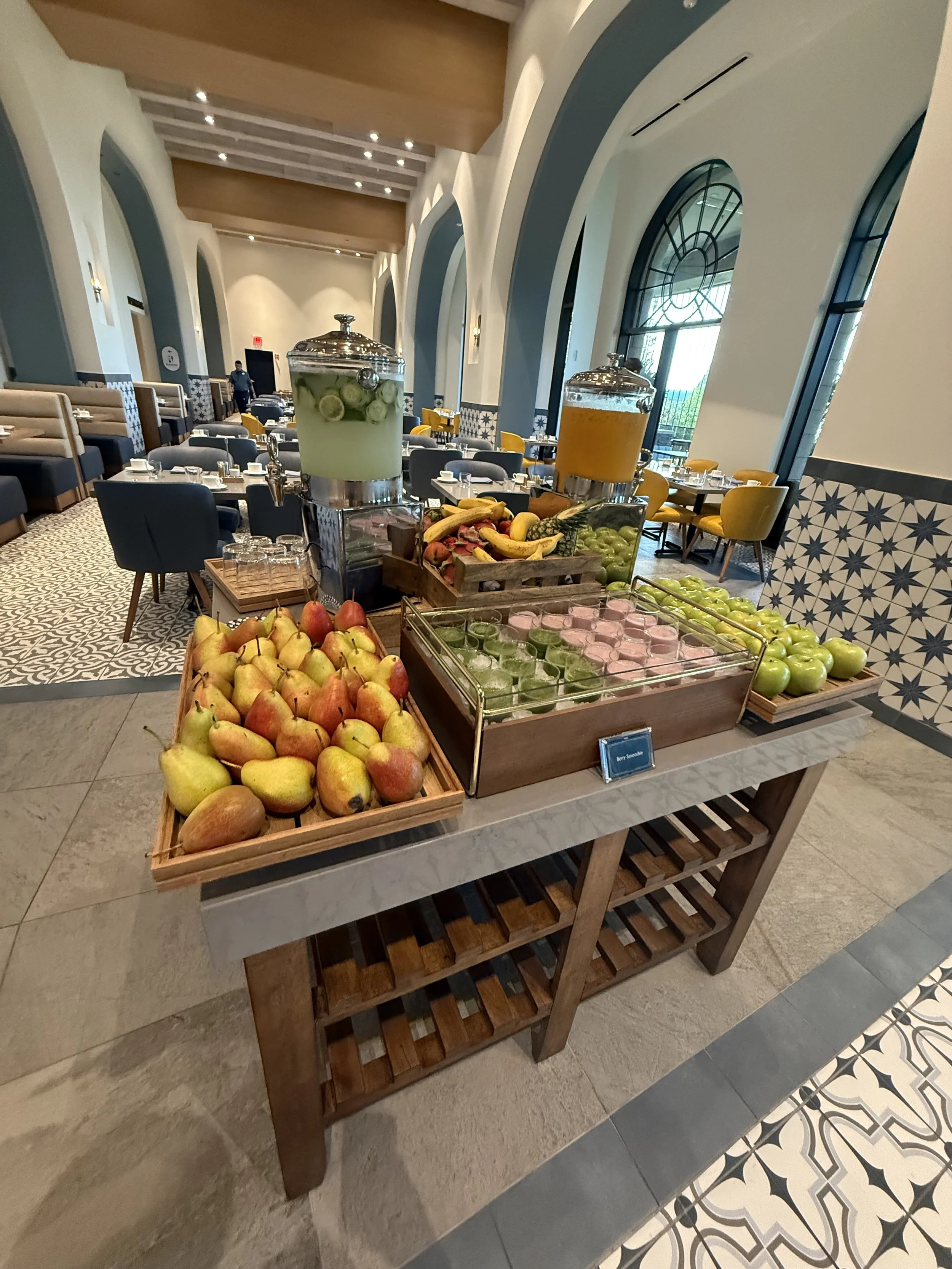 A fruit and beverage station in a hotel restaurant with apples, bananas, grapes, and melons, along with lemon and orange infused water and citrus juice.