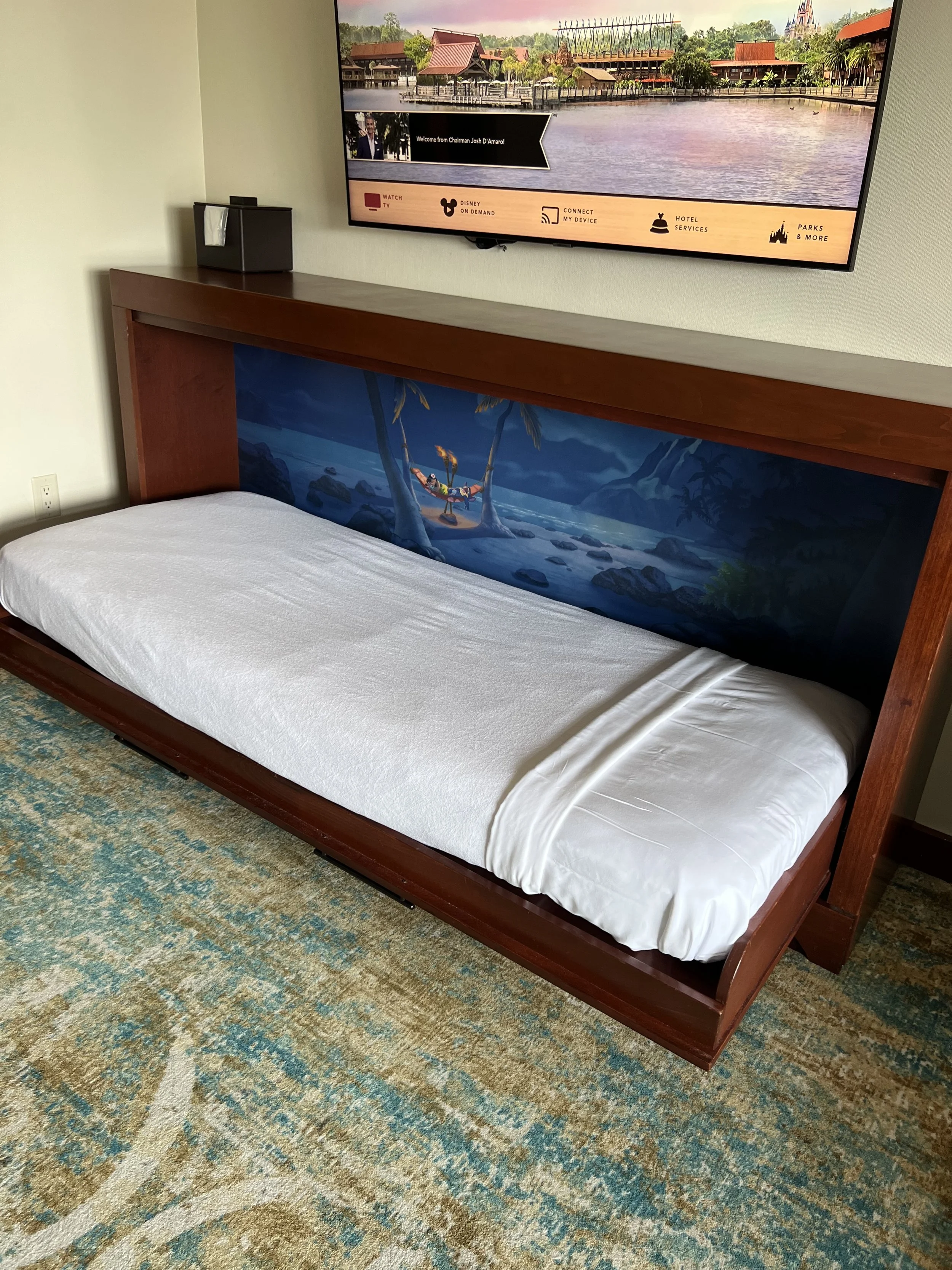 A hotel room with a single bed that has a decorative headboard depicting a beach scene with palm trees and a hammock. A large flat-screen TV is mounted on the wall above the bed, showing a landscape of a park or resort area near a body of water.