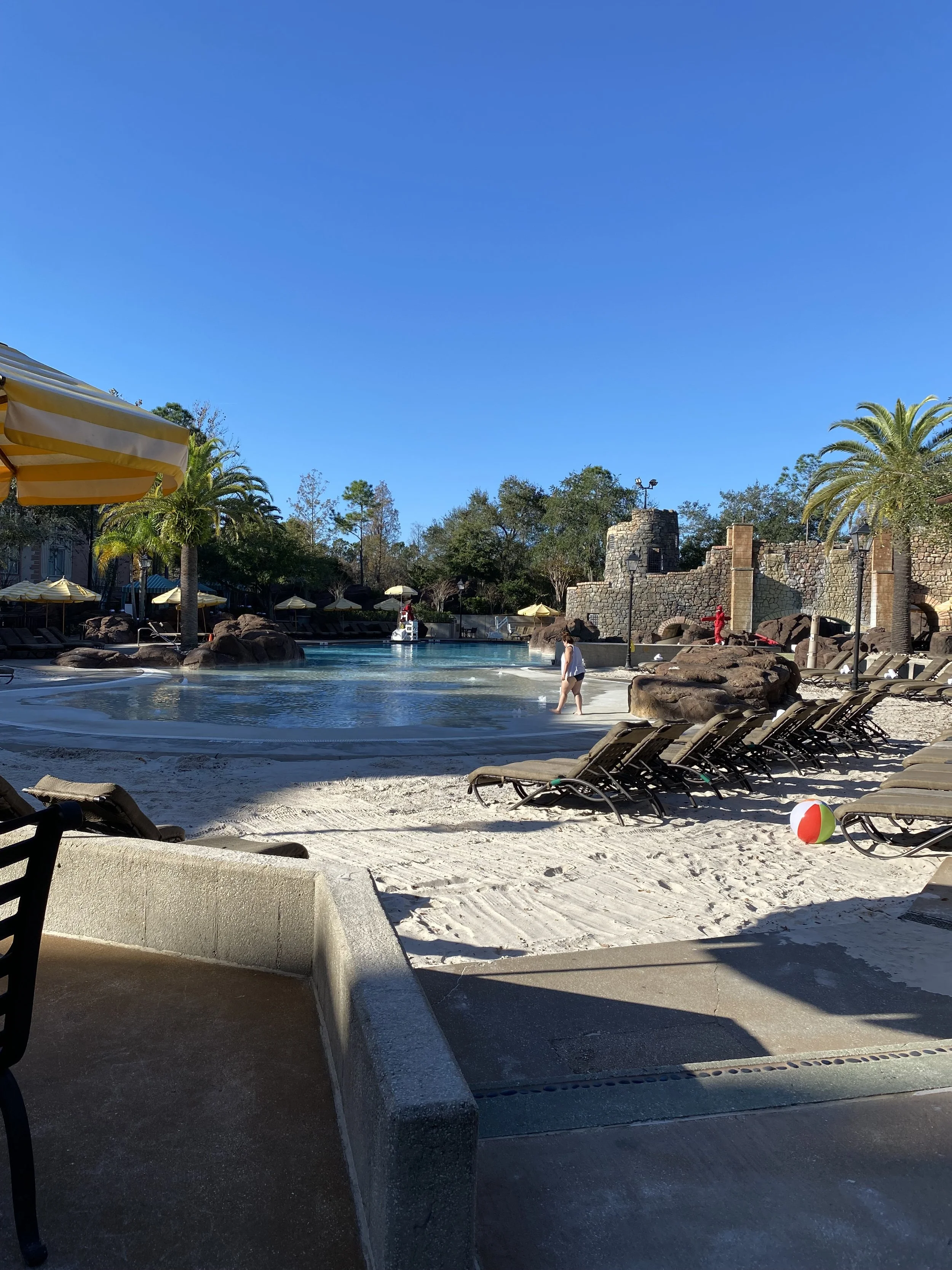 A swimming pool area with lounge chairs, a beach ball on the sand, tall palm trees, rocky features, and an arched stone structure under a clear blue sky.