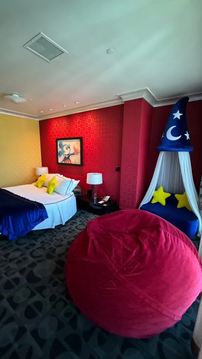 A hotel room with a bed, nightstand, lamps, and a large red bean bag chair. There is a wall with a portrait of a woman, and a whimsical blue wizard hat-shaped canopy over a seating area with yellow star pillows.