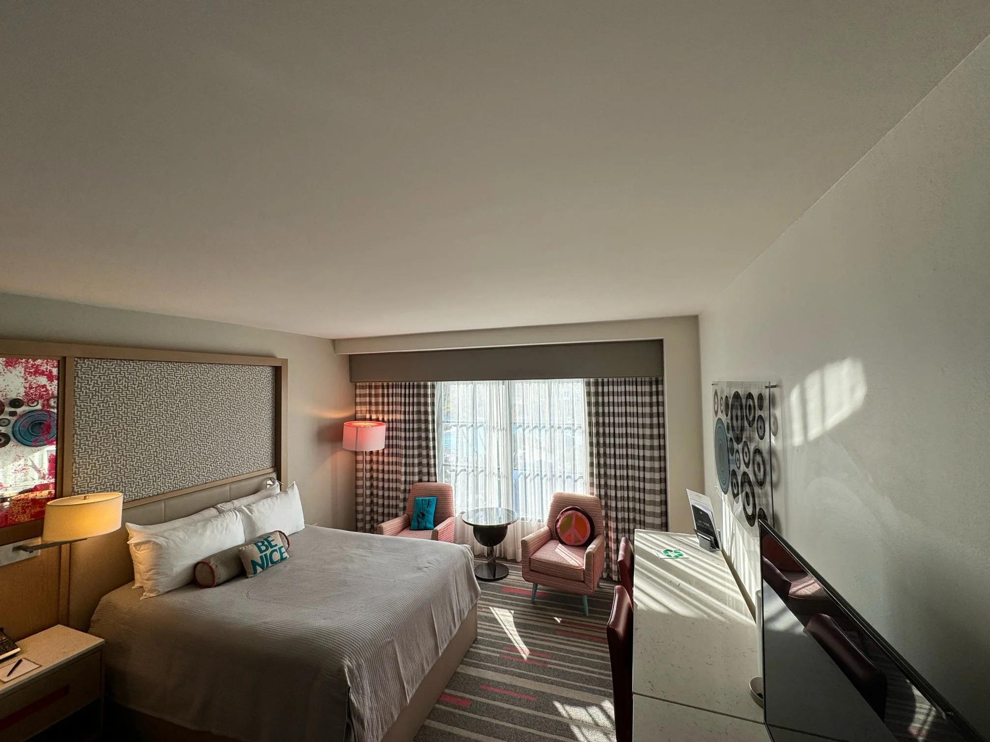 Interior of a modern hotel room with a bed, two pink armchairs, a small table, a desk, and a TV, with patterned curtains and artwork on the walls.
