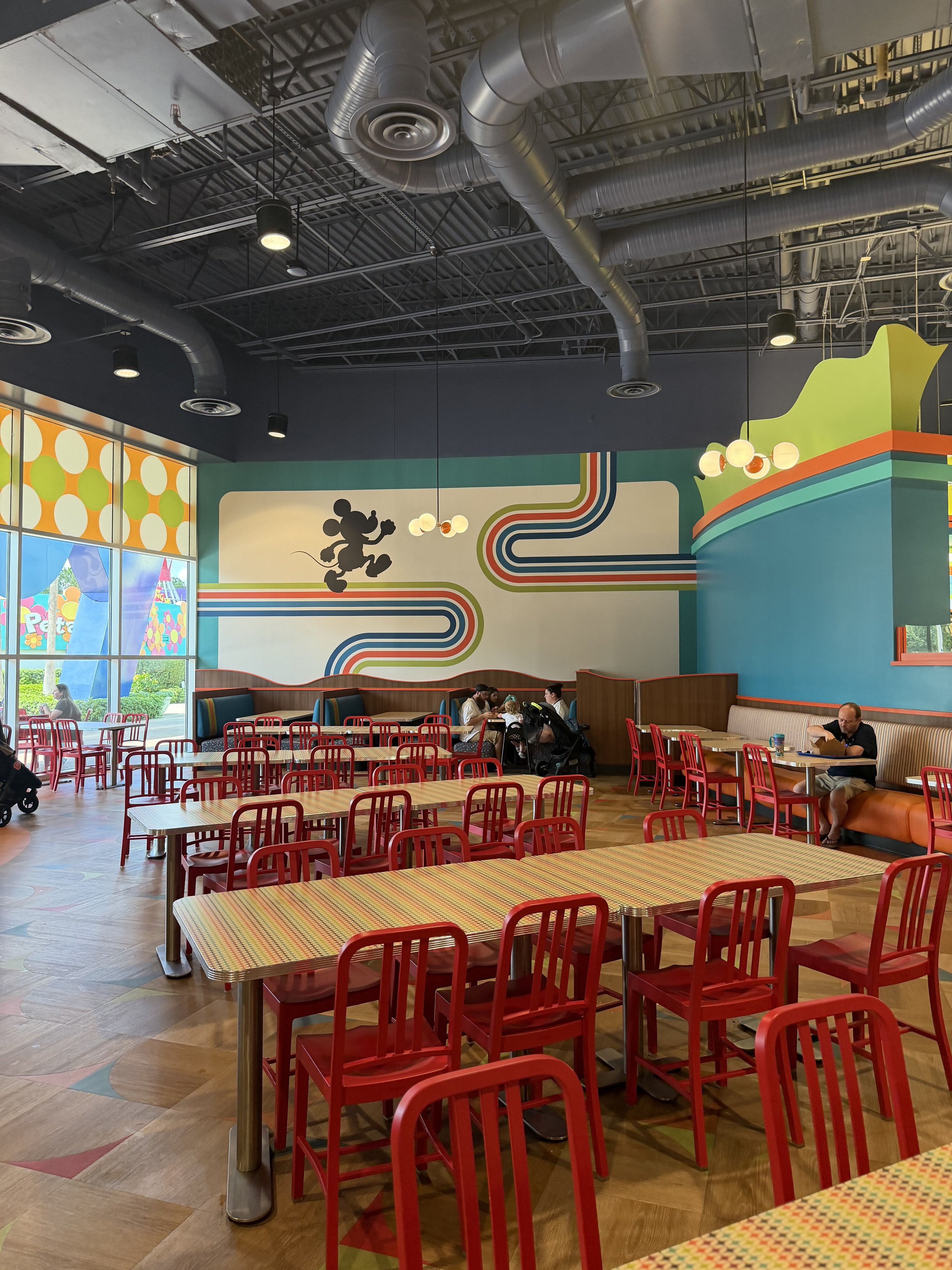 The image shows a colorful, Disney-themed restaurant interior with red chairs and tables, a mural of Mickey Mouse on the back wall, and some patrons sitting and eating.