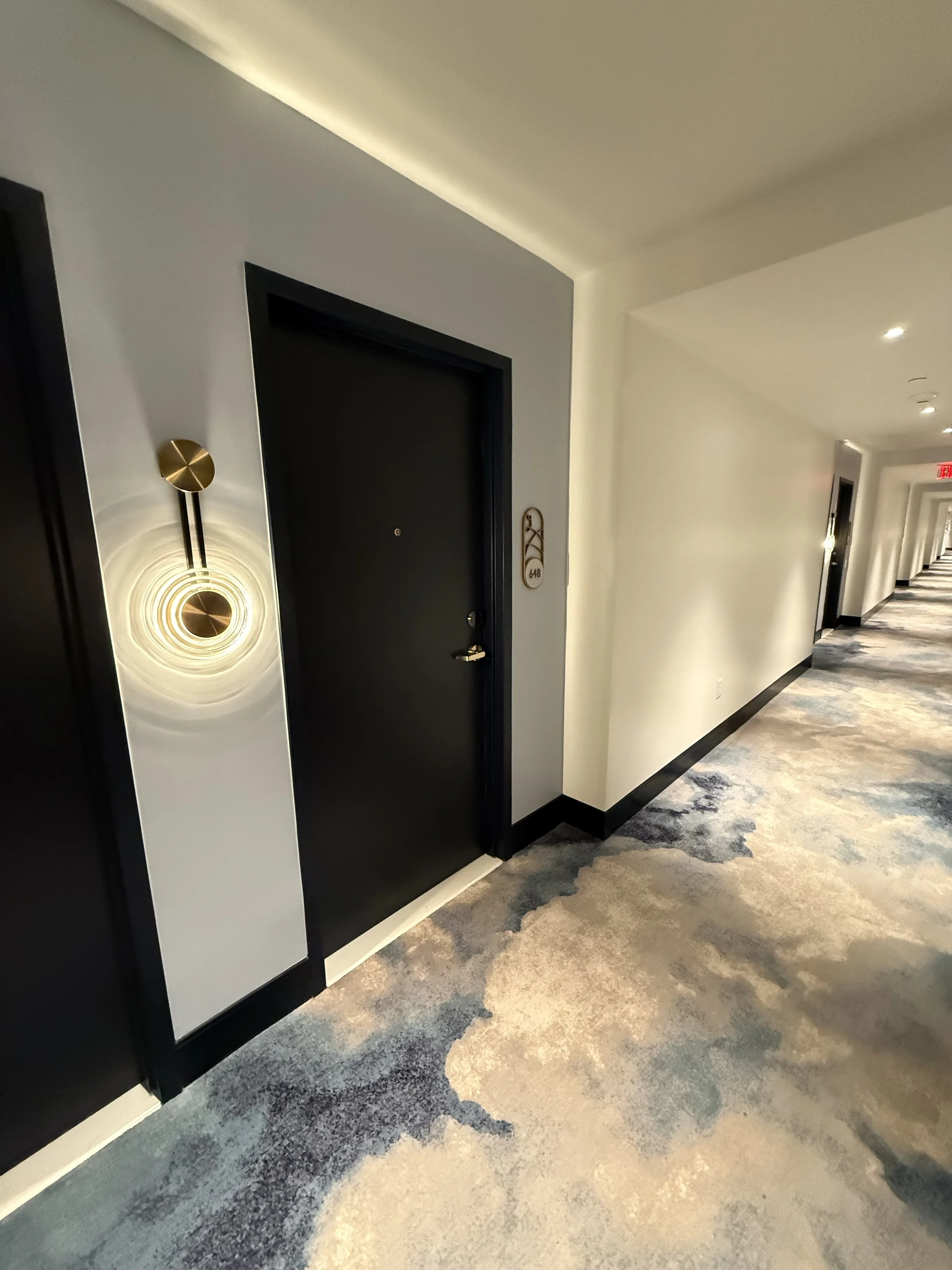 Hotel corridor with beige and blue patterned carpet, black doors on the left, illuminated wall sconces, and a black door with a room number 648.