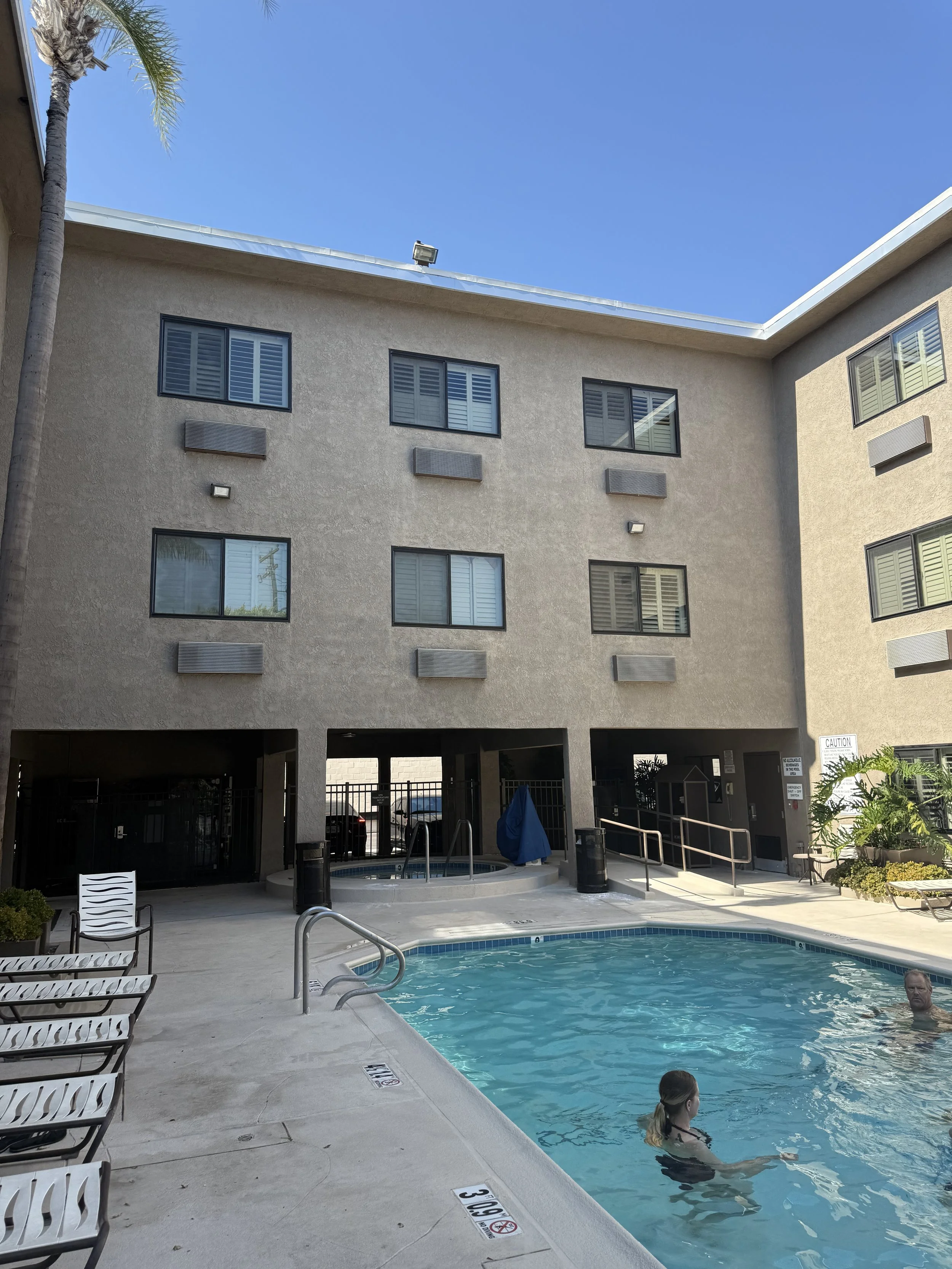 Outdoor pool area with two people swimming, surrounded by lounge chairs, with a multi-story beige apartment building and a clear blue sky in the background.