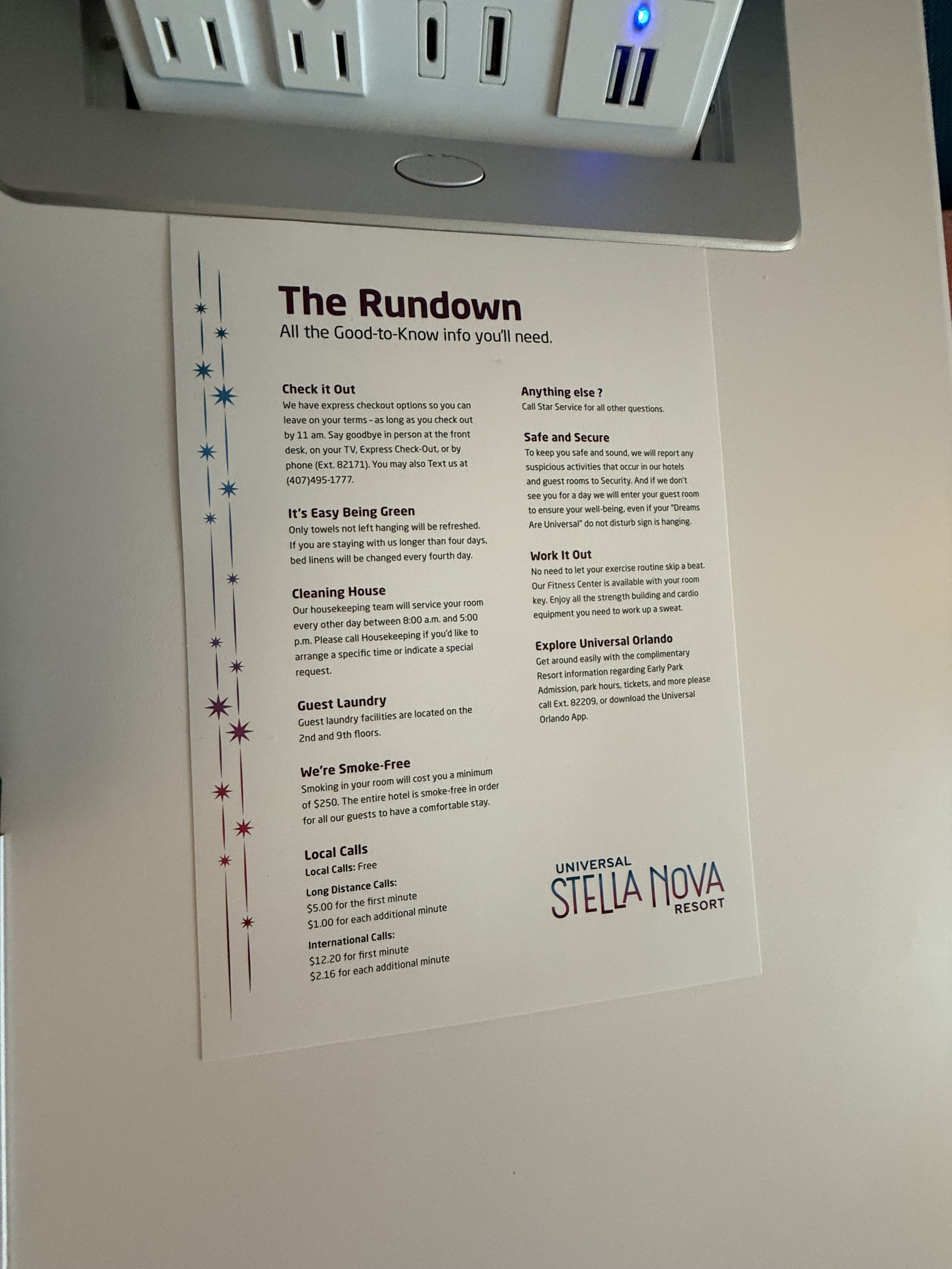 A printed informational sheet titled 'The Rundown' from Universal Stella Nova Resort, attached to a white surface near an electrical outlet with several plugs. The sheet provides hotel information including checkout procedures, towel policy, housekee
