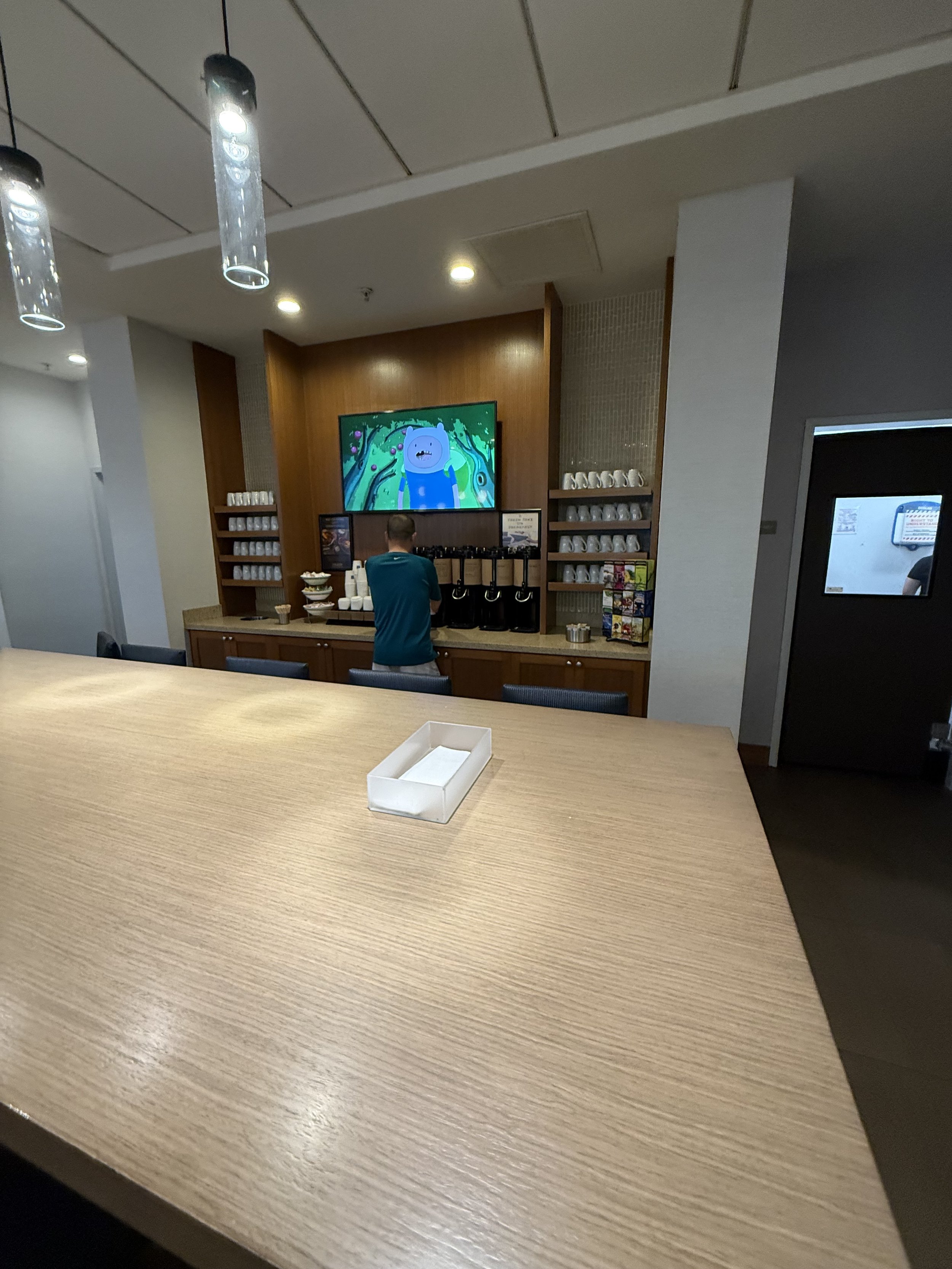 A hotel or cafe breakfast area with a wooden table in the foreground, a coffee station with a man in a teal shirt preparing coffee, and a large screen TV displaying cartoon characters in the background.