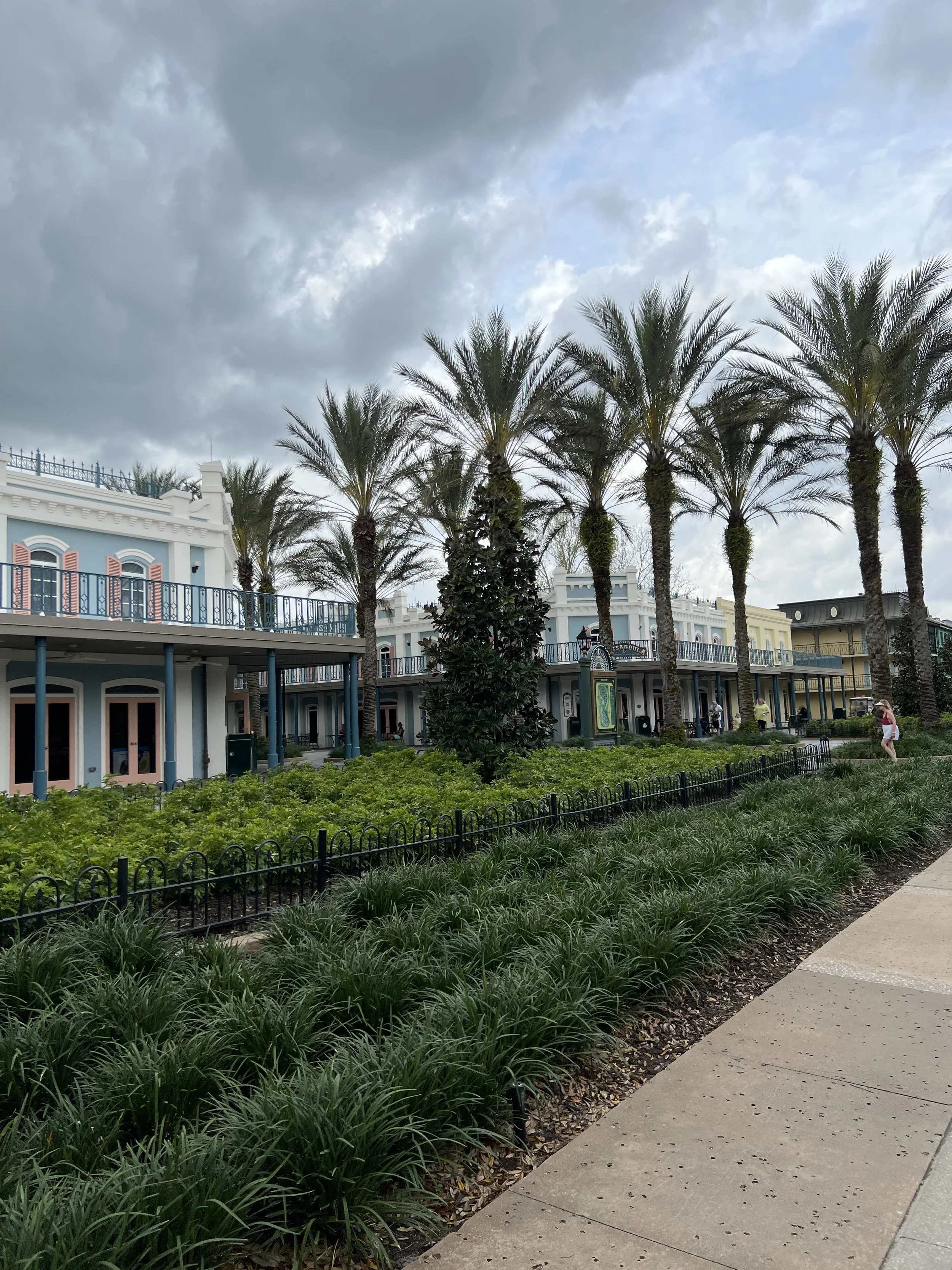 Colorful buildings with palm trees, green bushes, and a sidewalk on an overcast day.