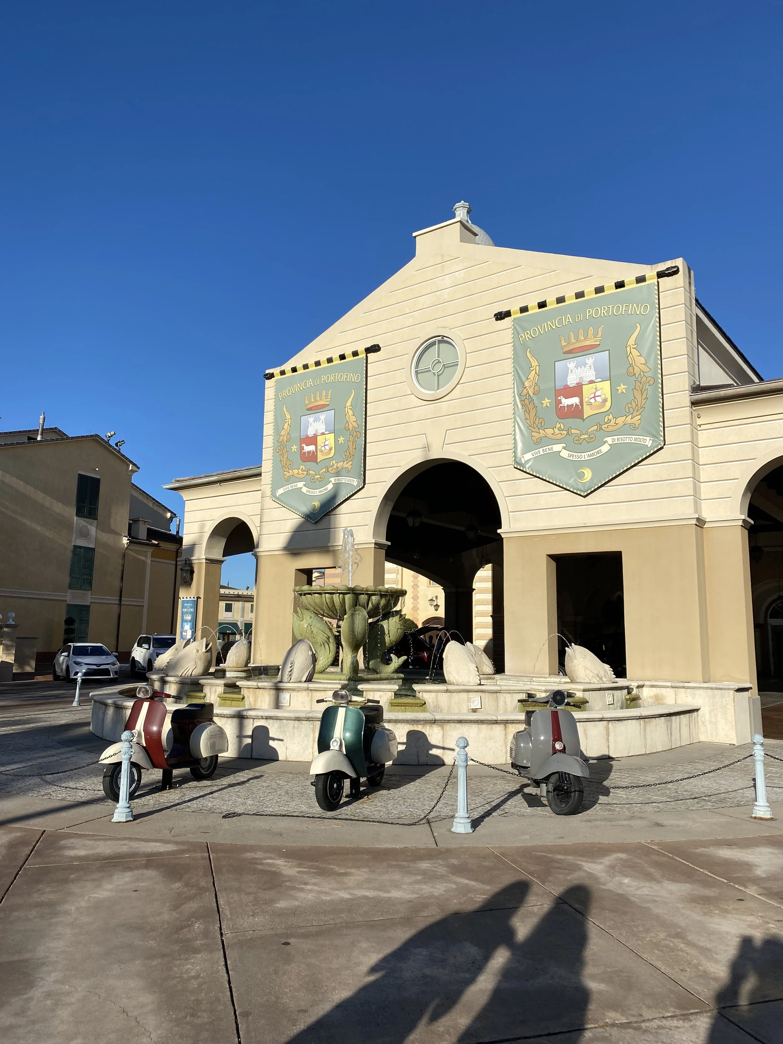 A beige building with an arched entrance and two banners displaying the crest of Portofino, Italy. A fountain with a large green clam shell sculpture and several dolphin statues is in front. Three vintage scooters are parked nearby on a paved area, a