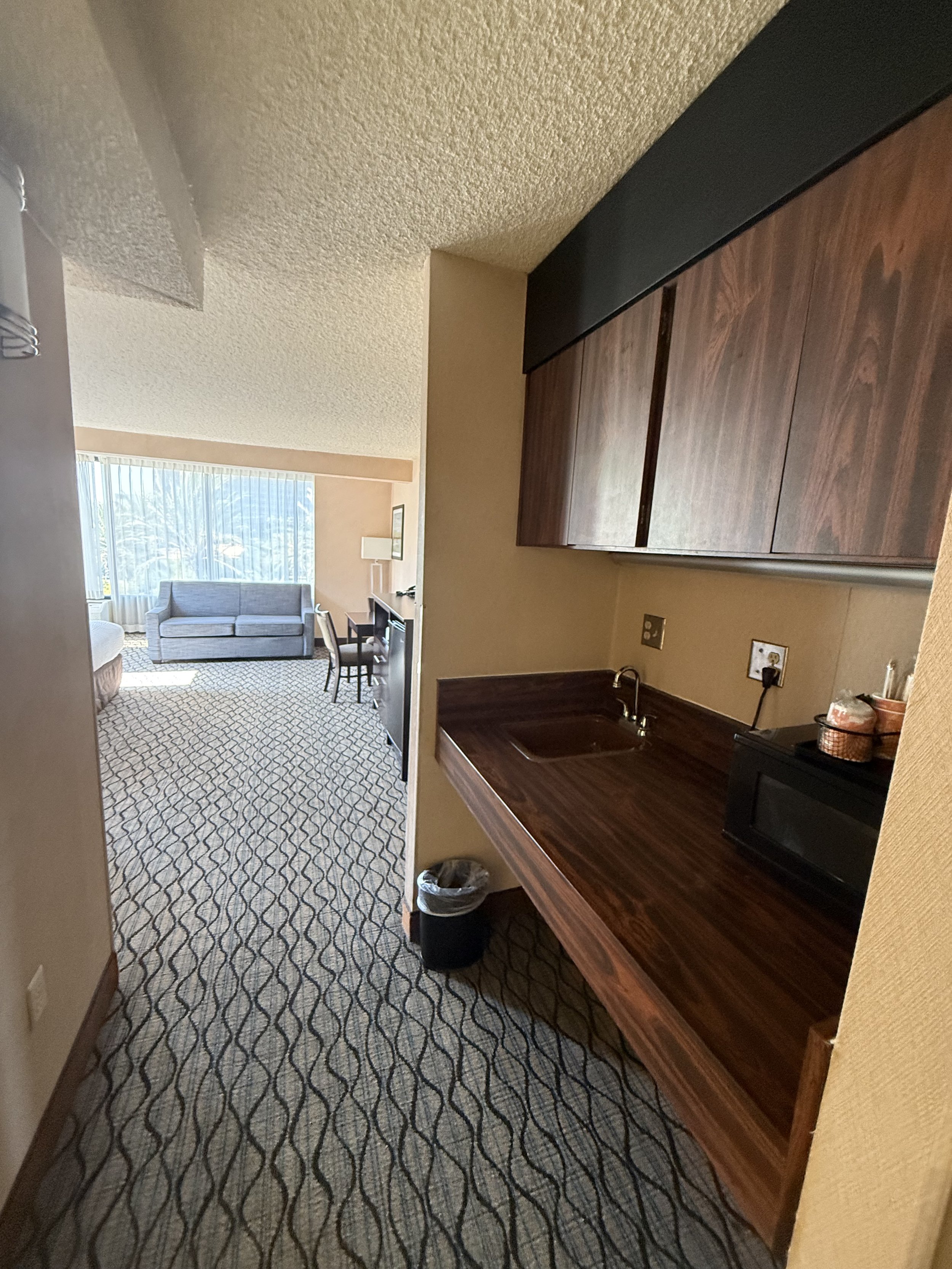 View of a hotel suite with a kitchenette area, wooden cabinets, a small sink, black microwave, and a trash bin, overlooking a living room with a sofa, chair, and large window with vertical blinds.