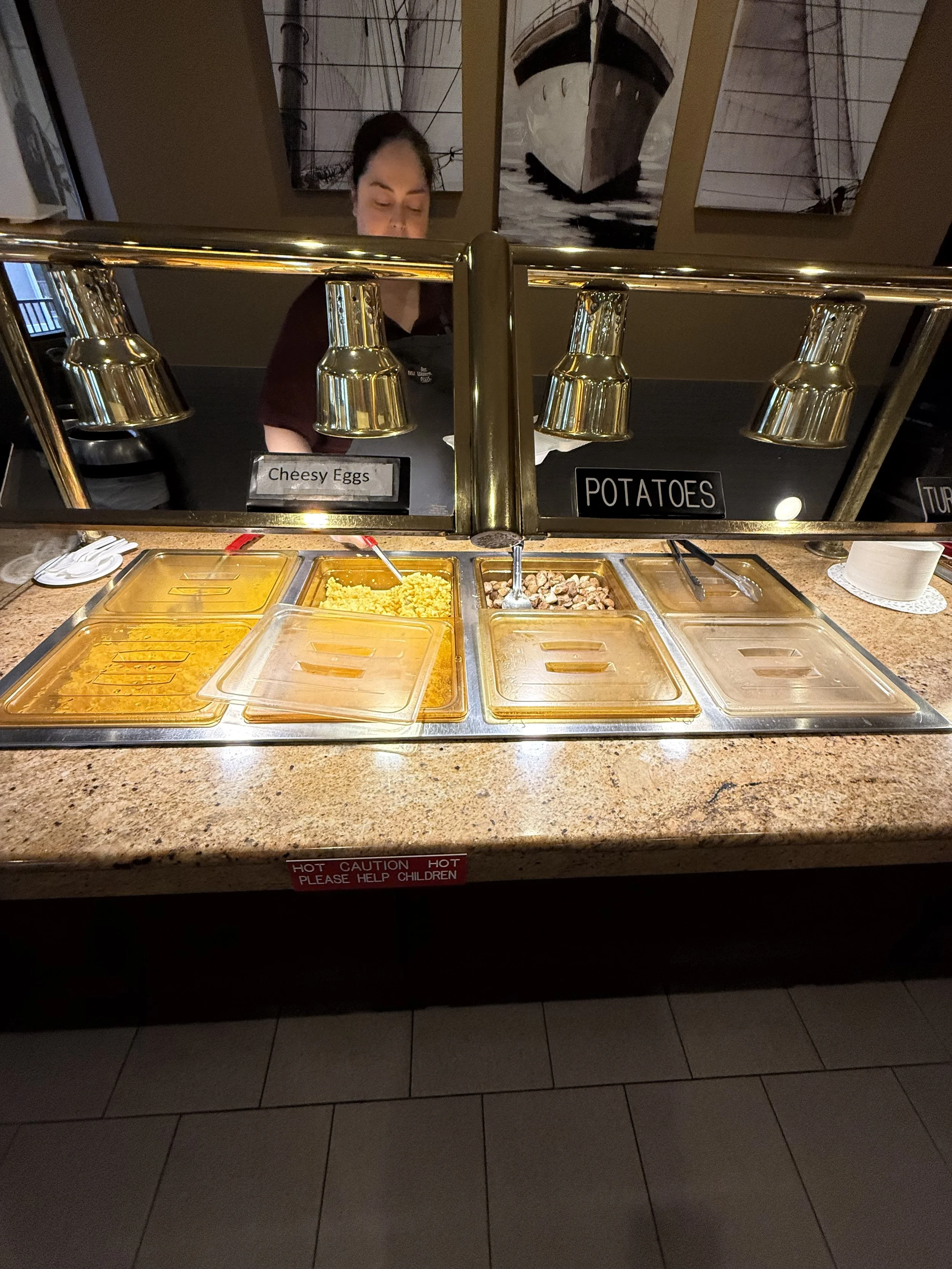 Buffet station with trays labeled 'Cheesy Eggs' and 'Potatoes', with a worker behind the counter and artwork of ships on the wall.