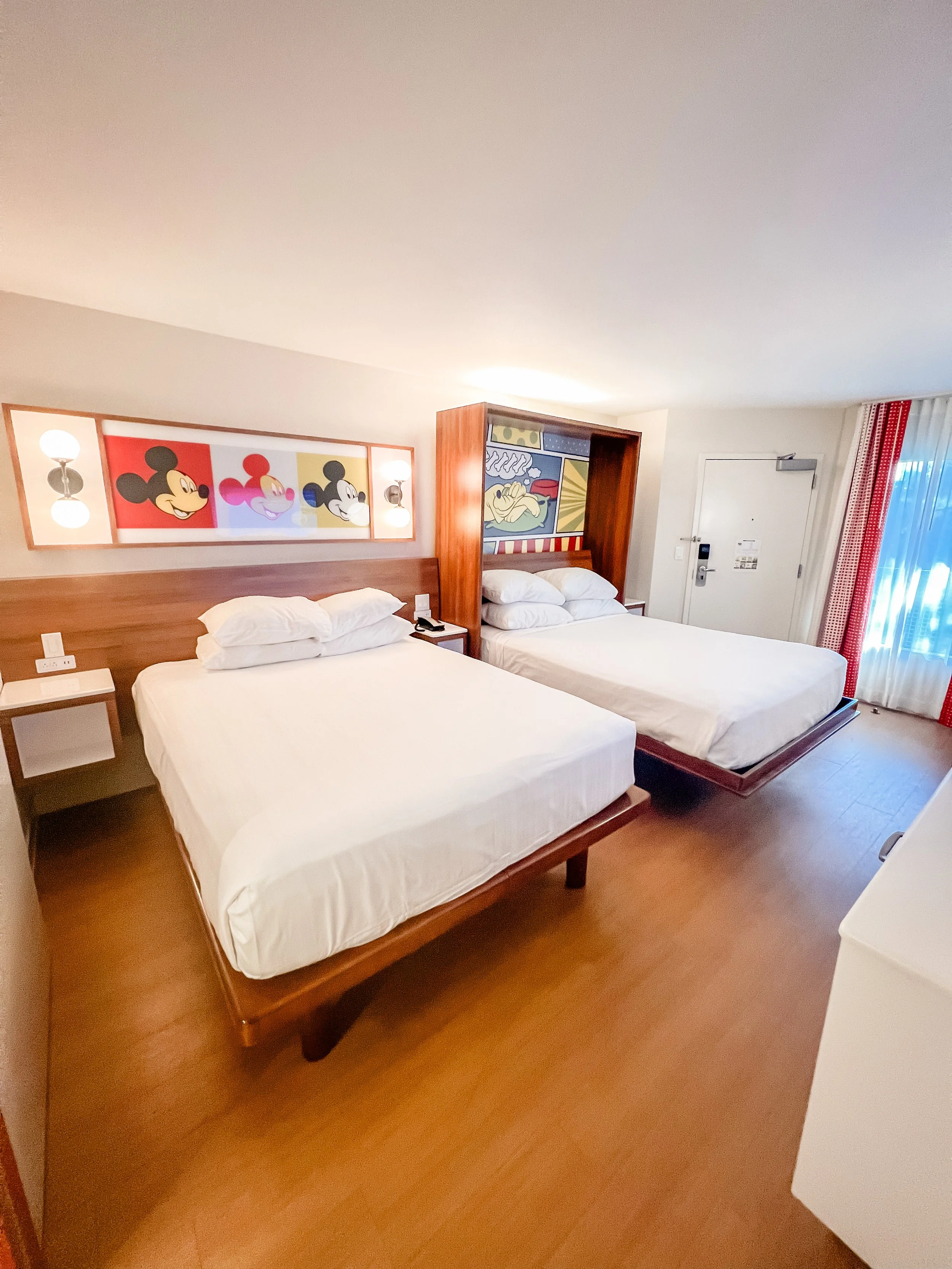 Hotel room with two beds, Mickey Mouse artwork above, and a colorful comic-style wall art panel.