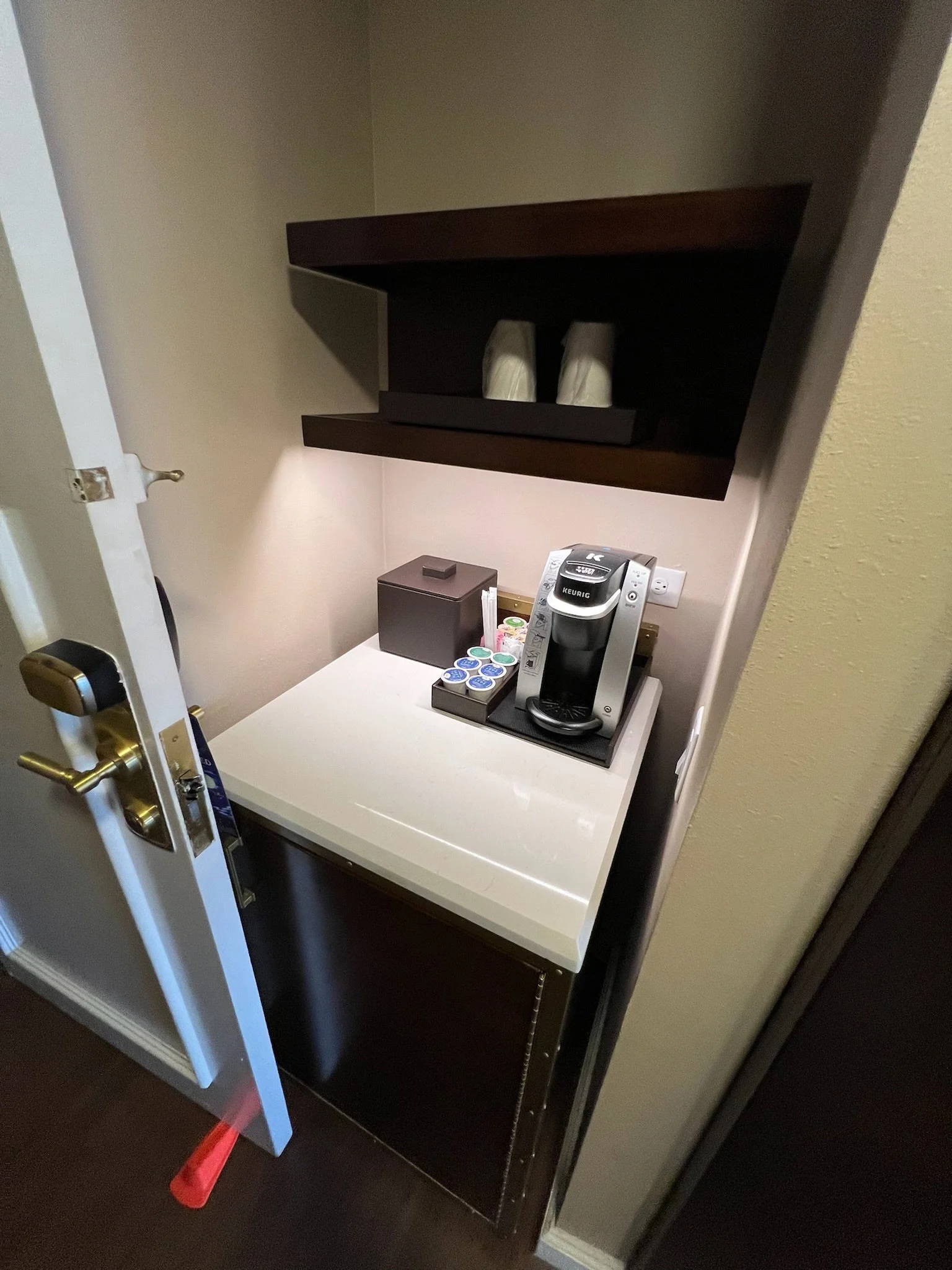 Small beverage station with a Keurig coffee maker on a white countertop, a box of coffee pods, a container with creamers, and a dark wooden shelf above holding two cups.