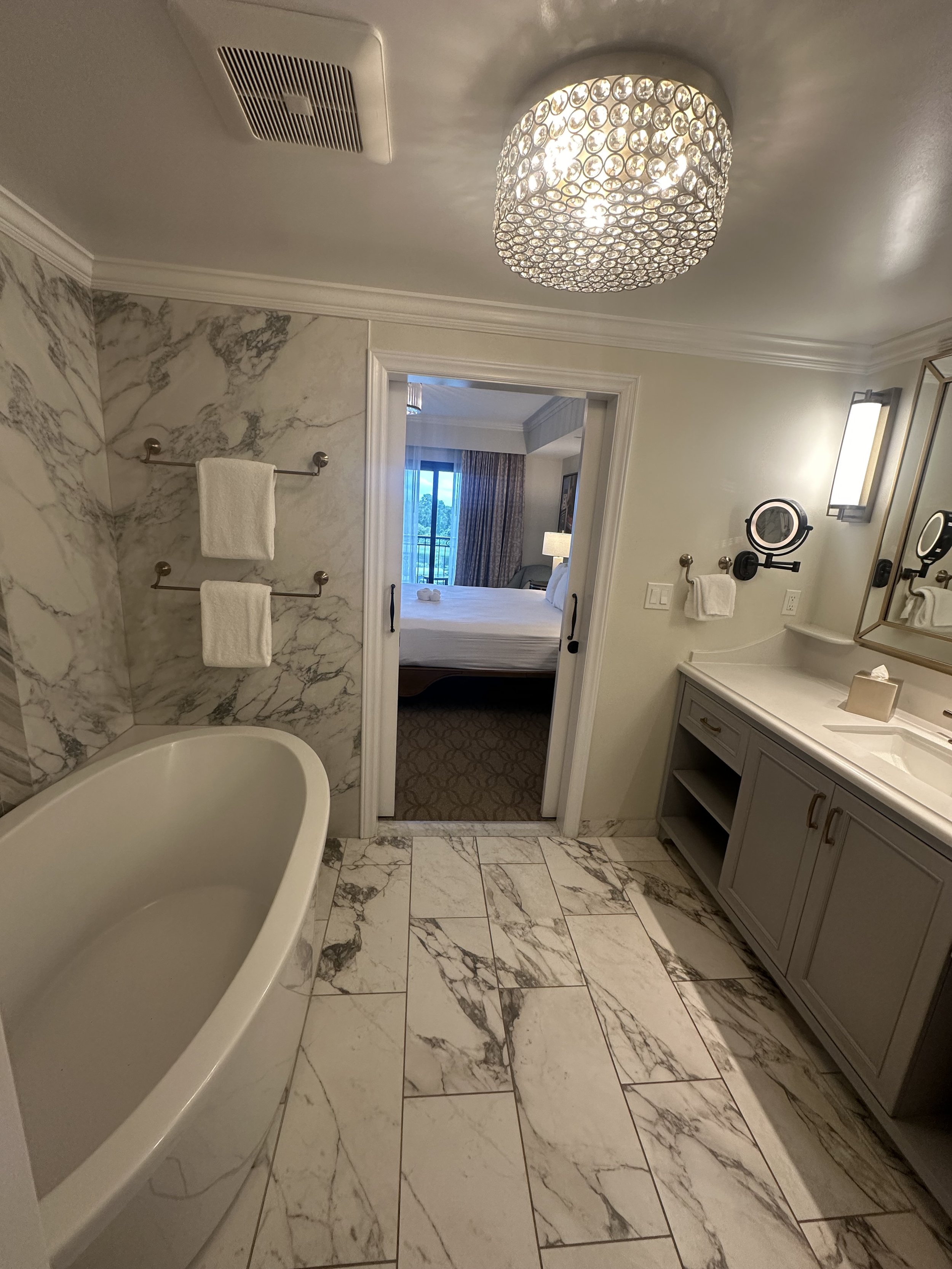 A luxurious bathroom with a marble bathtub, marble walls and floor, a bathroom vanity with a sink, mirror, and lighting, and a doorway leading to a bedroom with a bed and window.