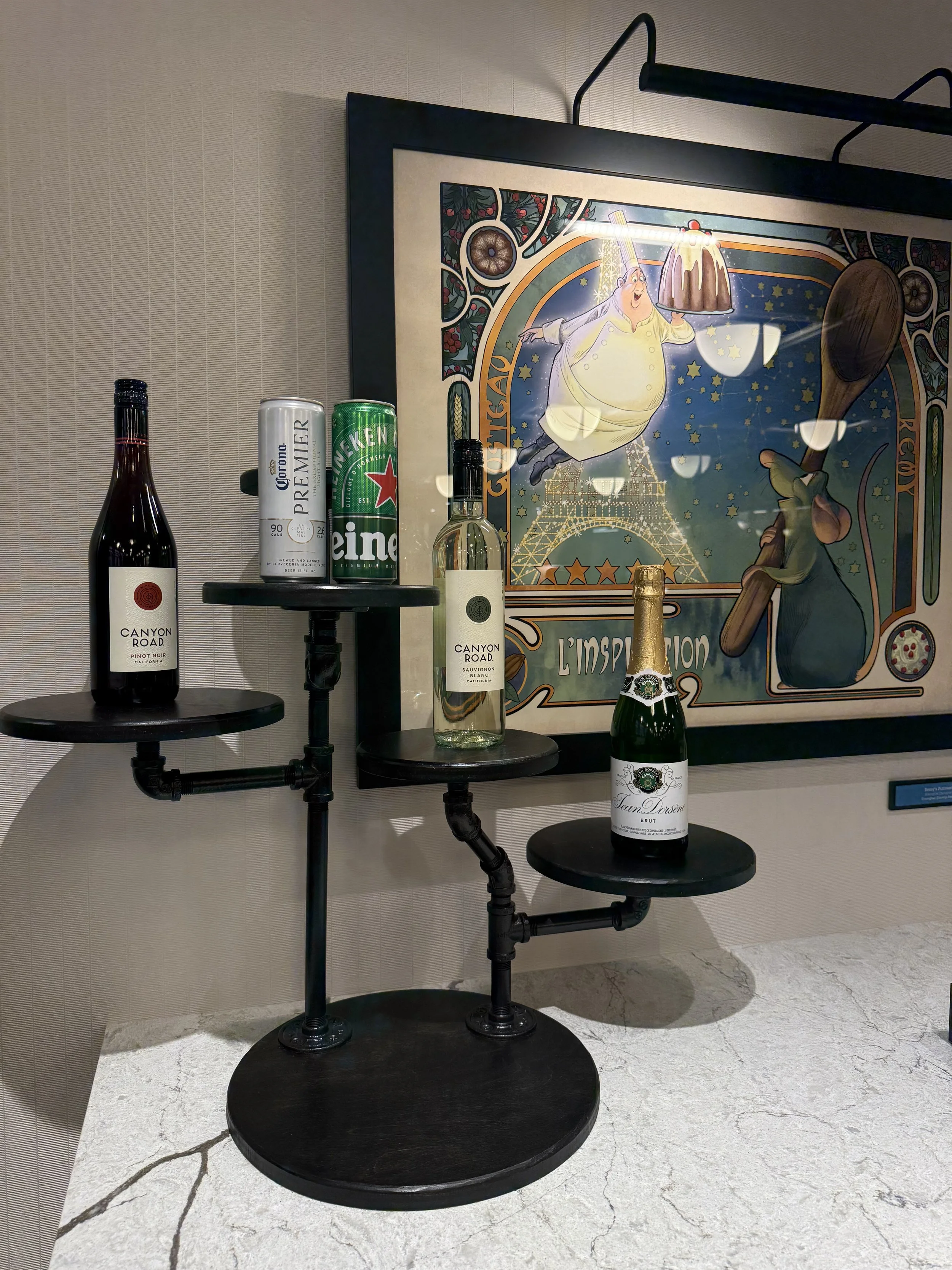 A black, multi-tiered pipe table with bottles of wine and a can of beer on top, set against a framed colorful artwork of a person in a chef's outfit and the Eiffel Tower.
