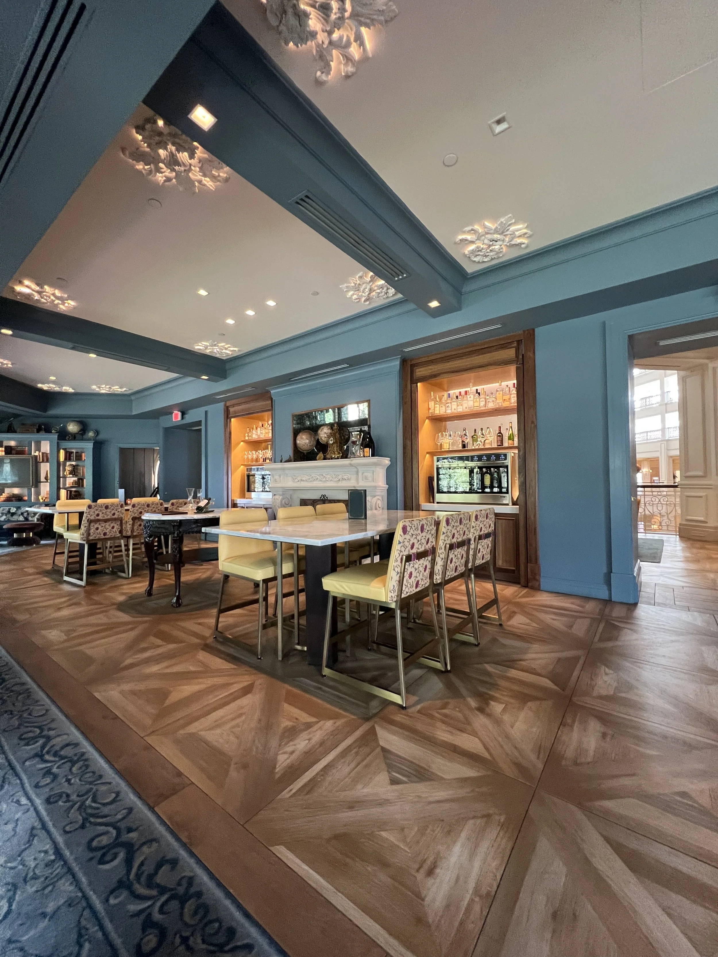 Elegant restaurant interior with blue walls, patterned wood flooring, and a variety of chairs around tables. There is a white fireplace with decorative items on top and open shelving displaying bottles and decorative pieces. The ceiling has ornate fl