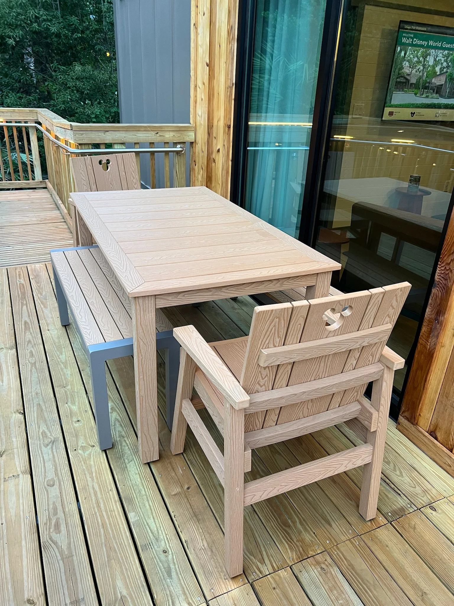 Wooden deck with a light wood table, a wooden chair, and a bench, outside a building with glass sliding doors.