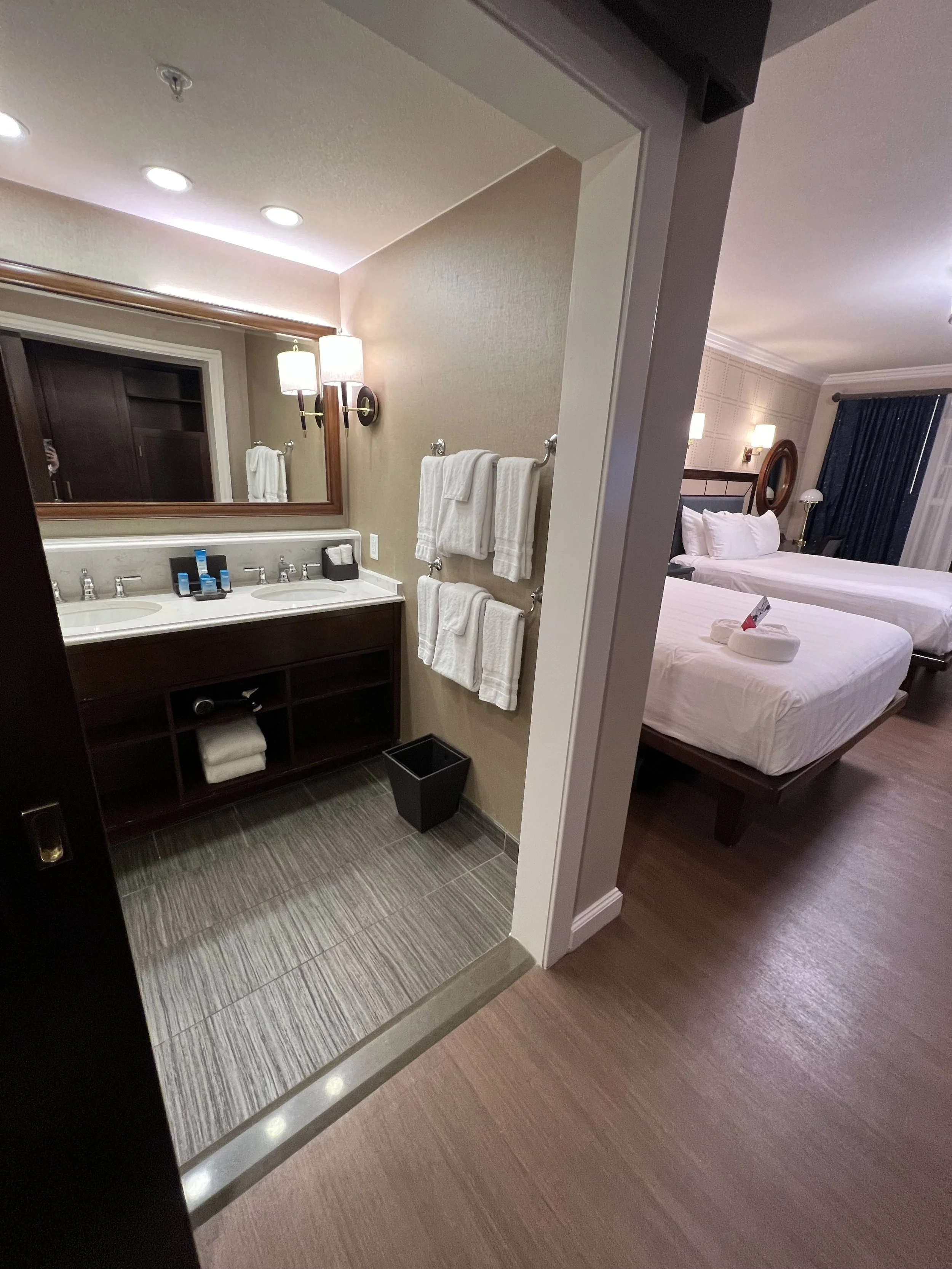 Hotel bathroom with double sink, mirror, towels, toiletries, and a trash can. Bedroom with two beds, pillows, a nightstand, lamp, and window with curtains.