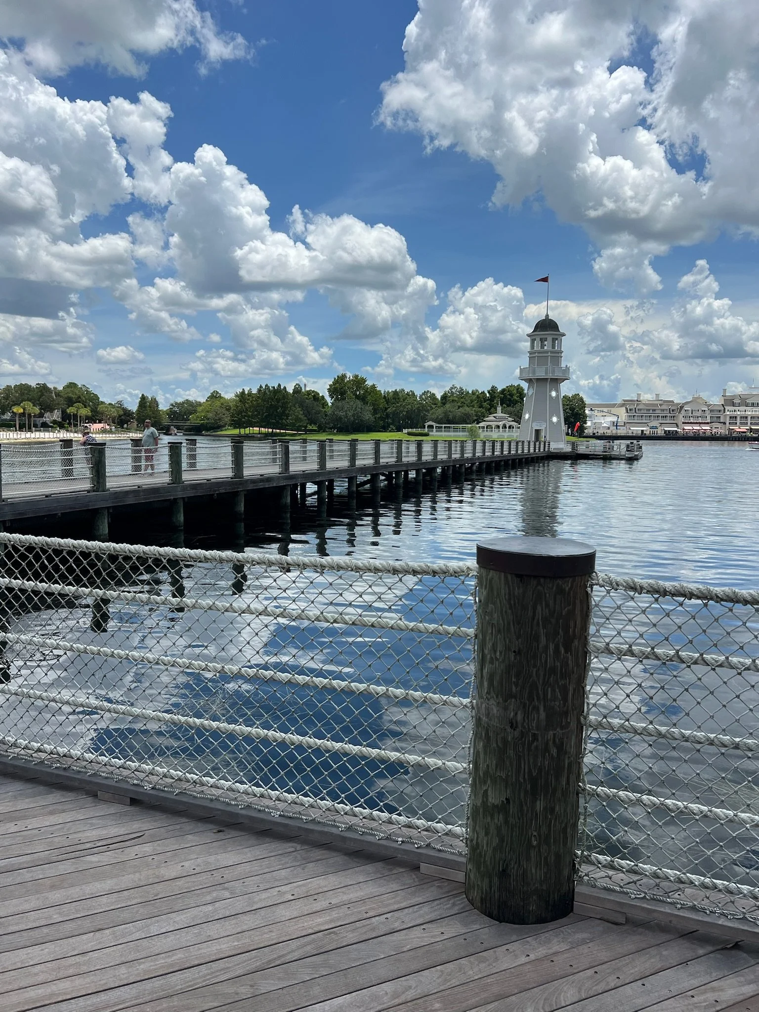 A wooden pier with a white safety fence extending over calm water. In the background, a white lighthouse with a flag on top stands near a park with trees. The sky is mostly blue with white, fluffy clouds.