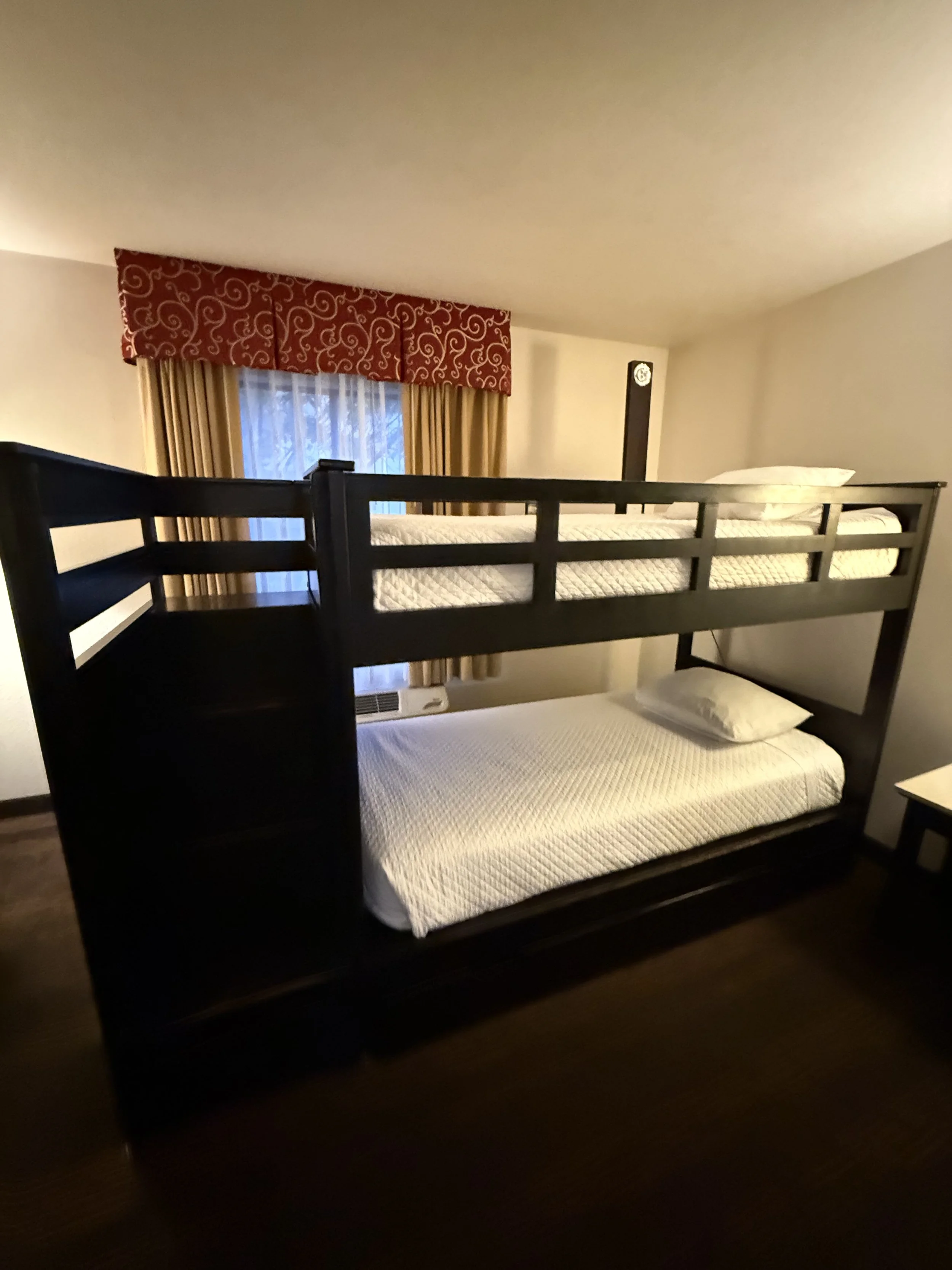 A room with a bunk bed, featuring a single bed on the bottom and a mattress on the top, against a wall with a window. The window has beige curtains and a red valance with white swirls.