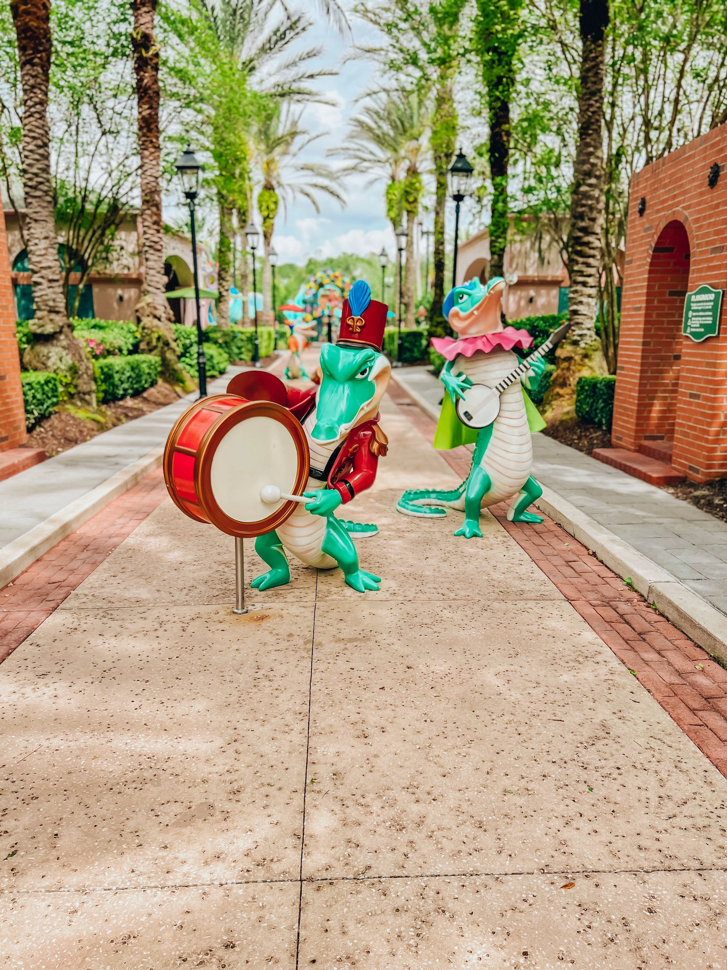 Colorful frog sculptures dressed as musicians, one playing a drum and the other playing a guitar, along a sidewalk in a lush, green outdoor setting.