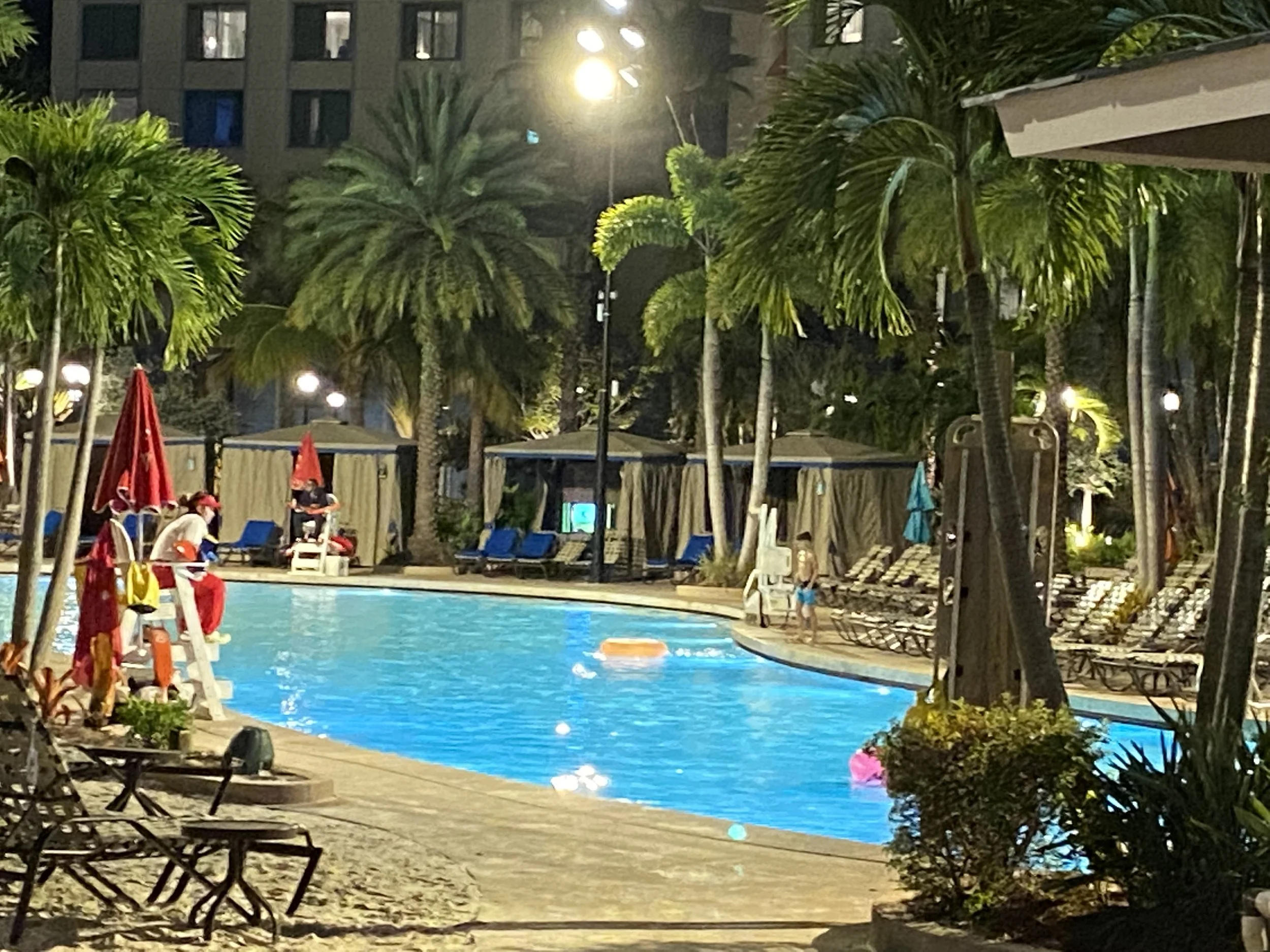 Night view of a swimming pool area at a resort with palm trees, lounge chairs, umbrellas, and people enjoying the pool, illuminated by outdoor lights.