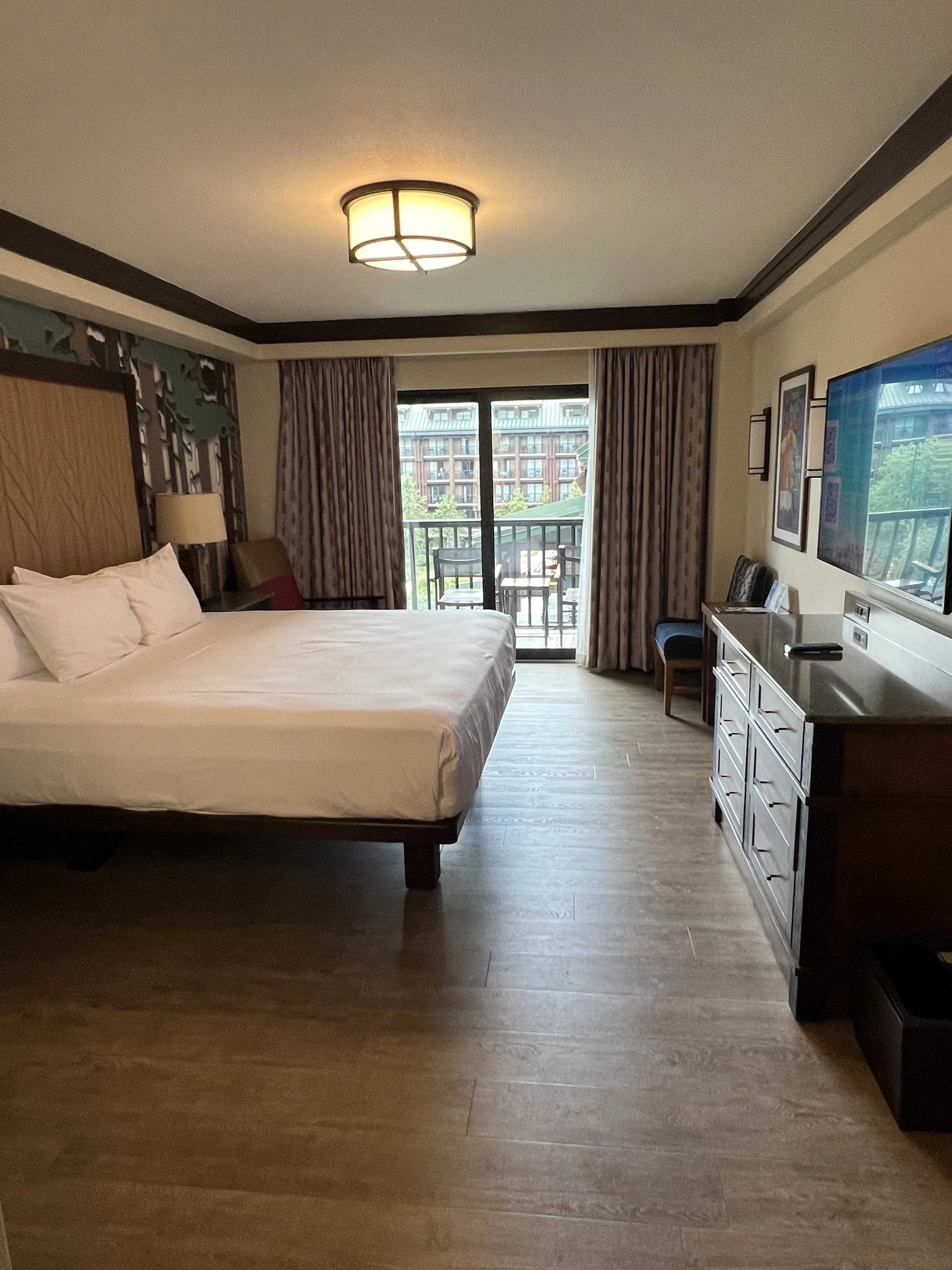 Hotel room with a large bed, a TV, desk, balcony with outdoor seating, and urban view outside.