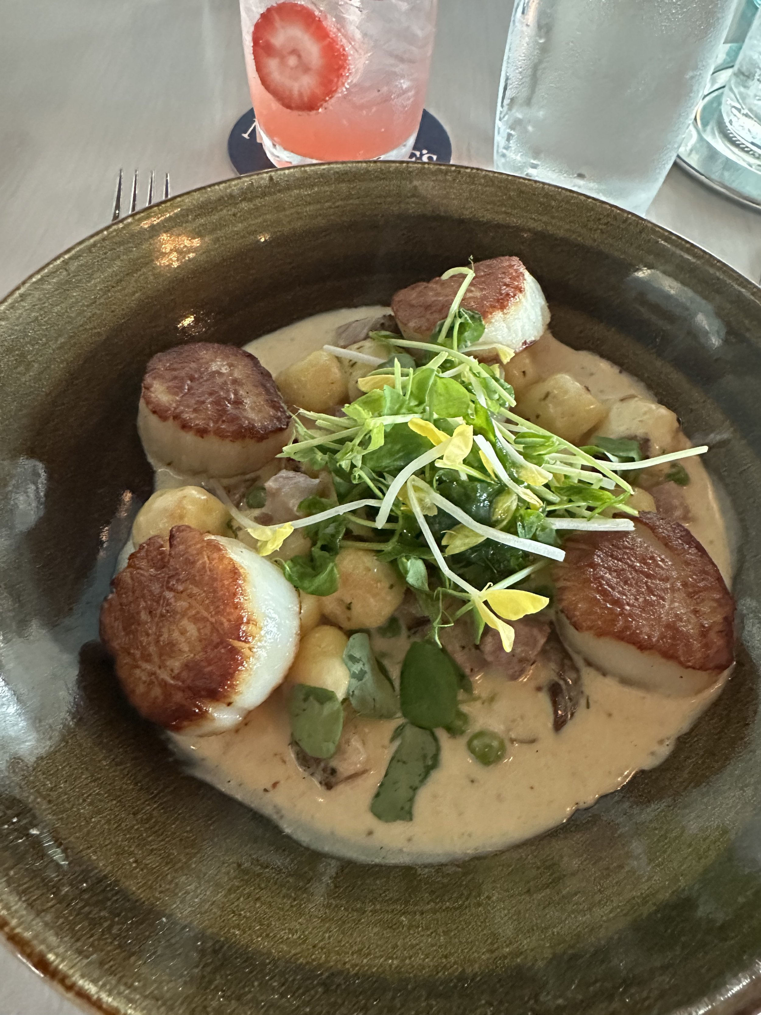 A bowl of cooked scallops served with a creamy sauce, garnished with microgreens, with a drink containing a strawberry and a lemon slice, and glasses of water in the background.
