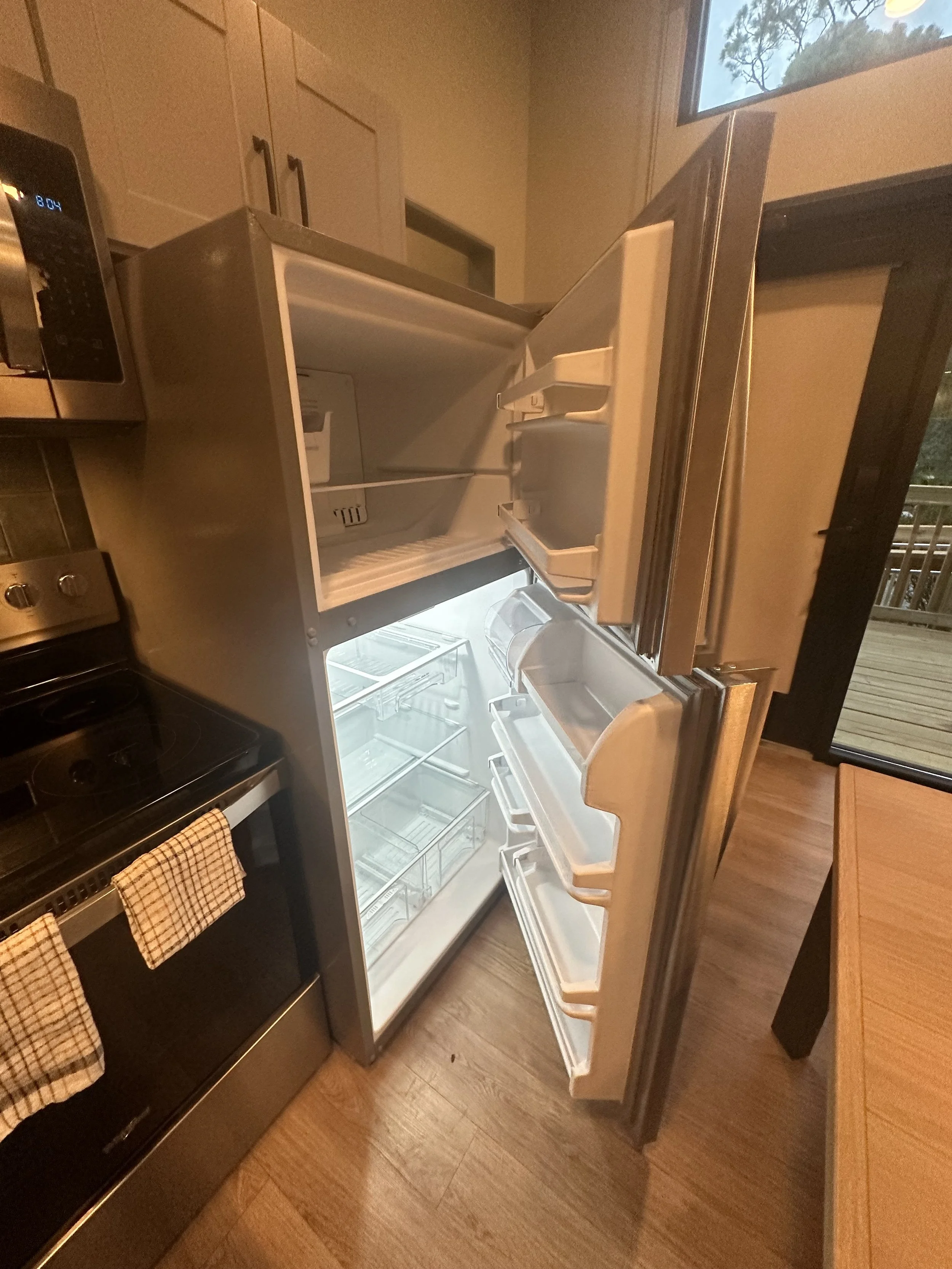 Open refrigerator with empty shelves in a kitchen with a microwave, stove, and wooden floor, and a sliding glass door leading outside.