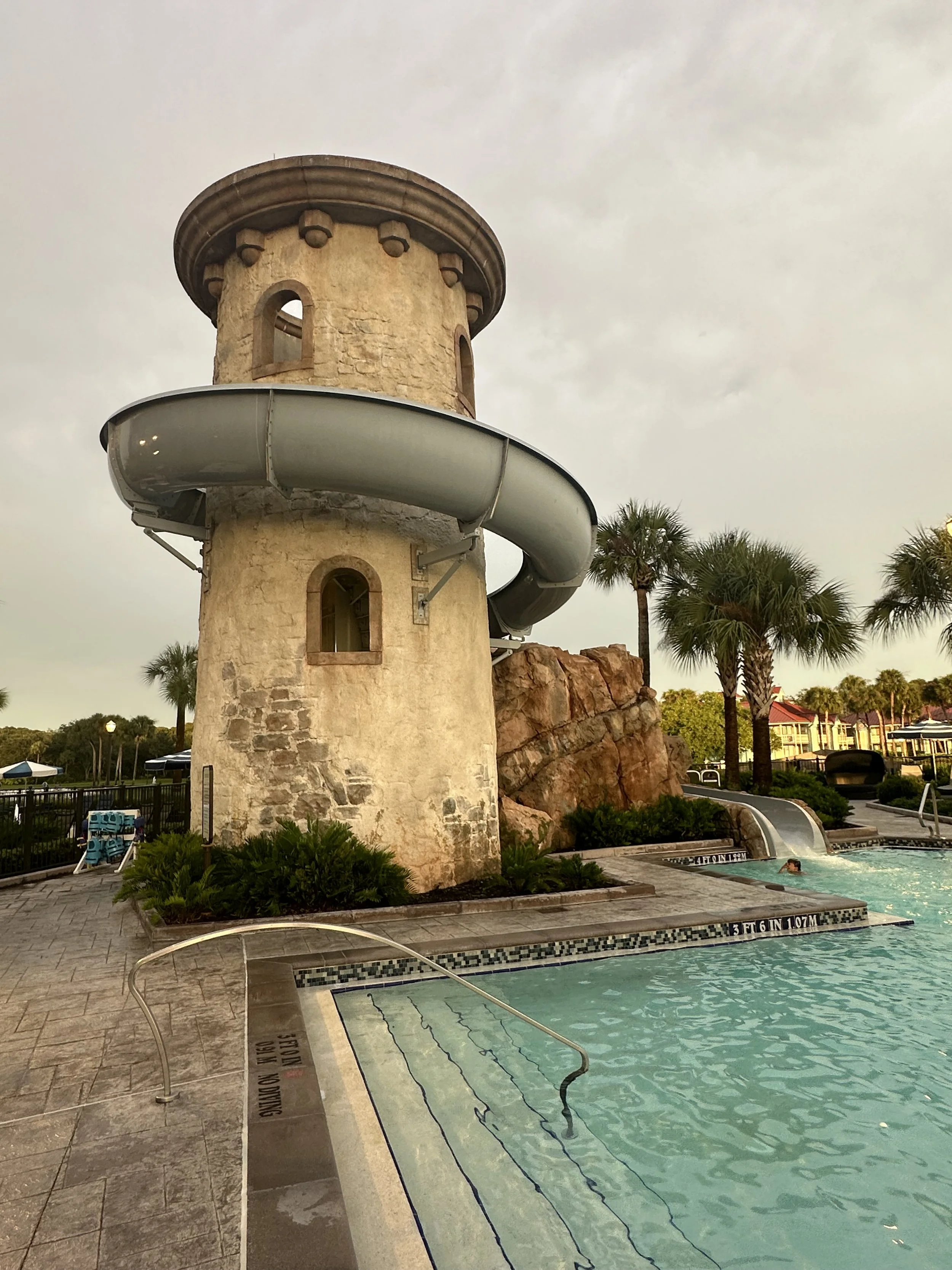 A water park with a tall stone tower and a large water slide wrapping around it. There is a pool at the base of the tower with a person swimming. Palm trees and buildings are visible in the background.