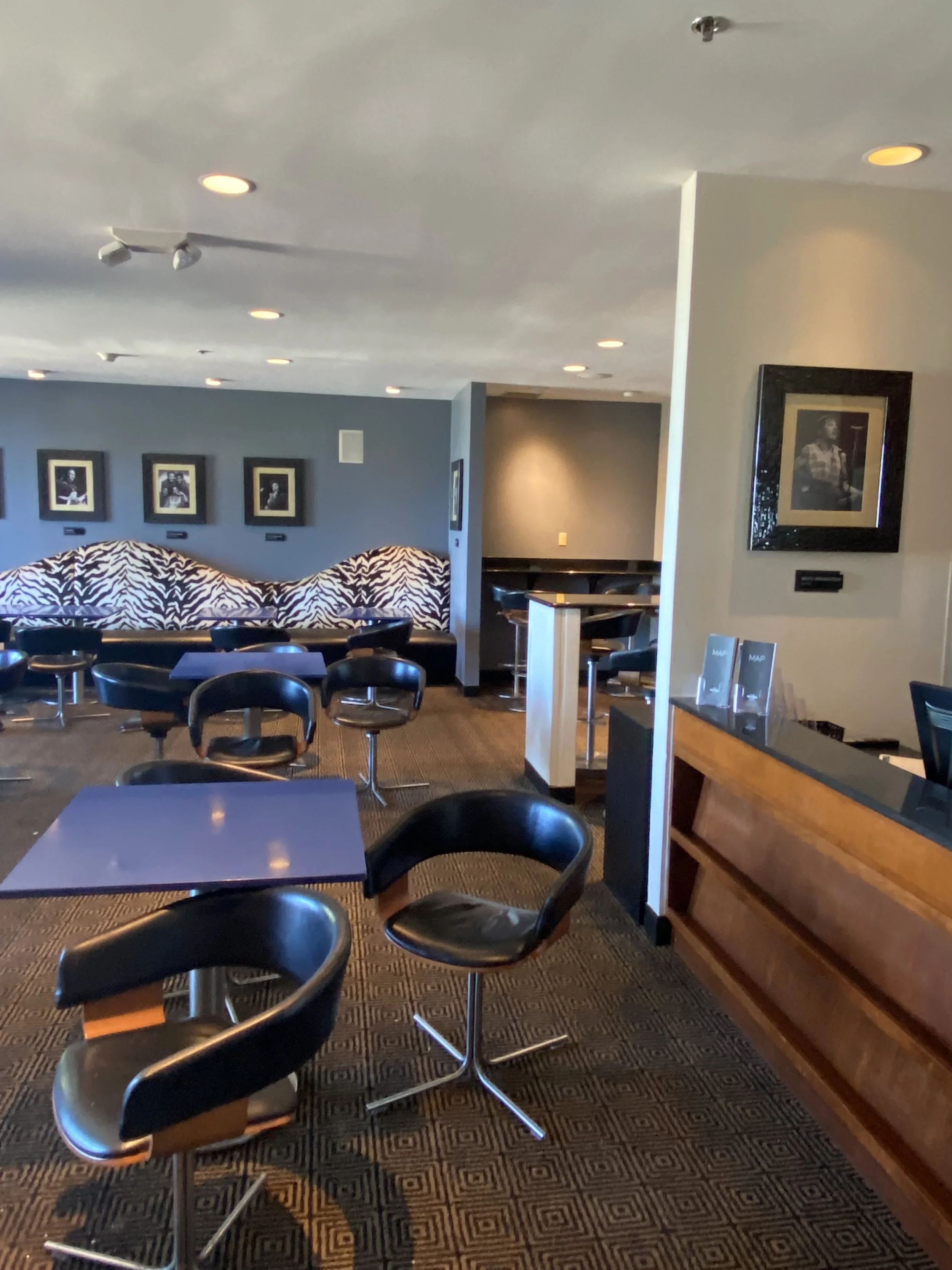 Interior of a bar or lounge with black swivel chairs, small tables, zebra print cushioned seats, framed black and white photographs on the walls, and a carpeted floor.