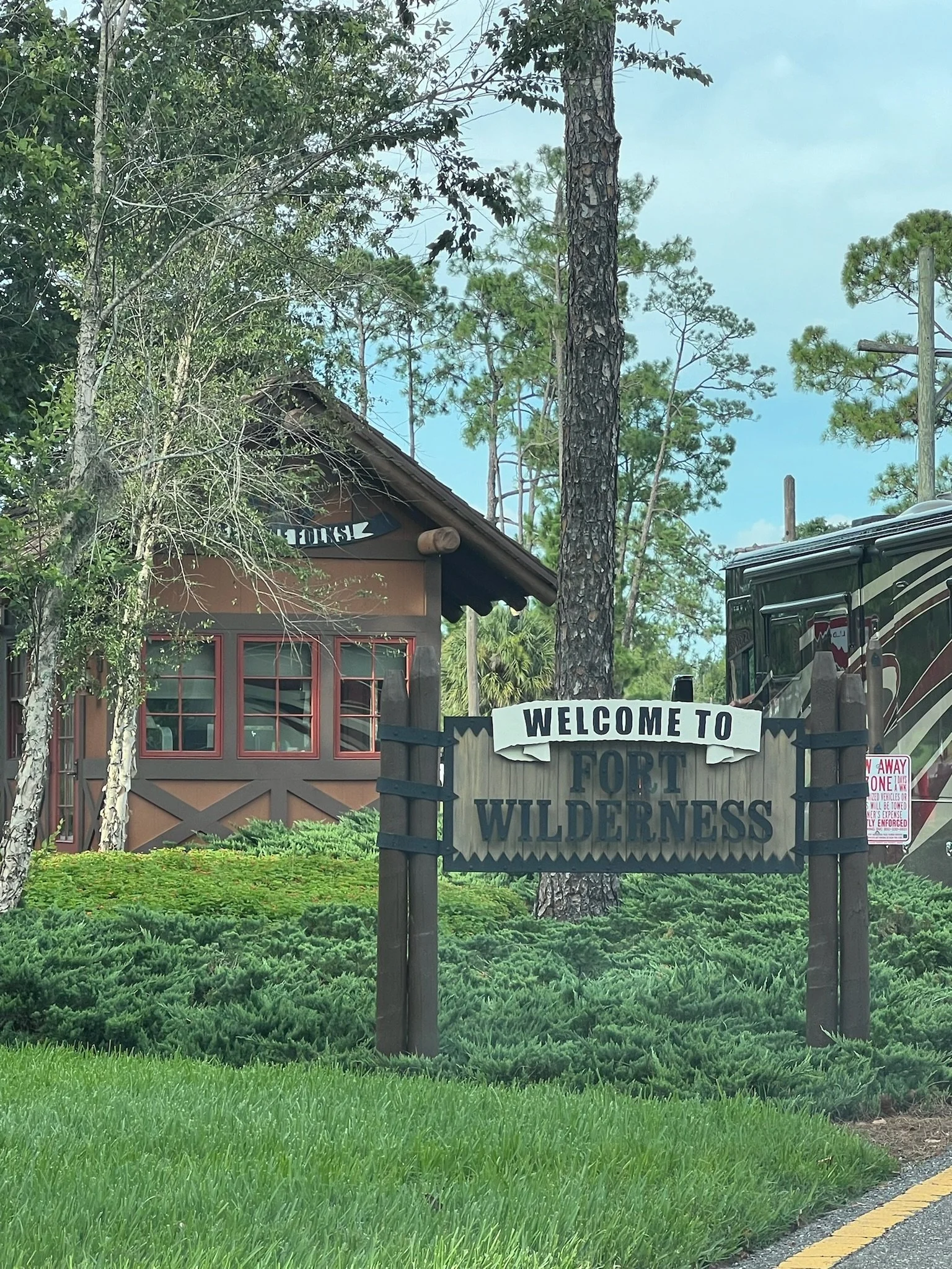 A sign reading 'Welcome to Fort Wilderness' in front of a cabin surrounded by greenery and trees, with a large RV parked nearby.