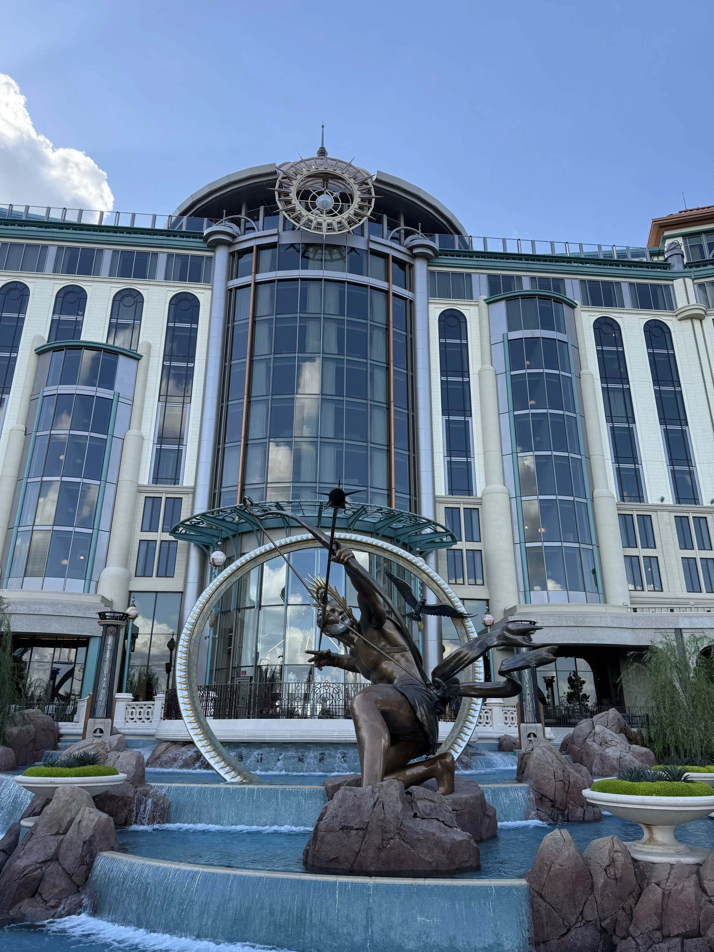 A large modern building with blue glass windows and a clock near the top. In front, there's a fountain with a bronze sculpture of a horse and a person holding a spear, surrounded by rocks and small plants.