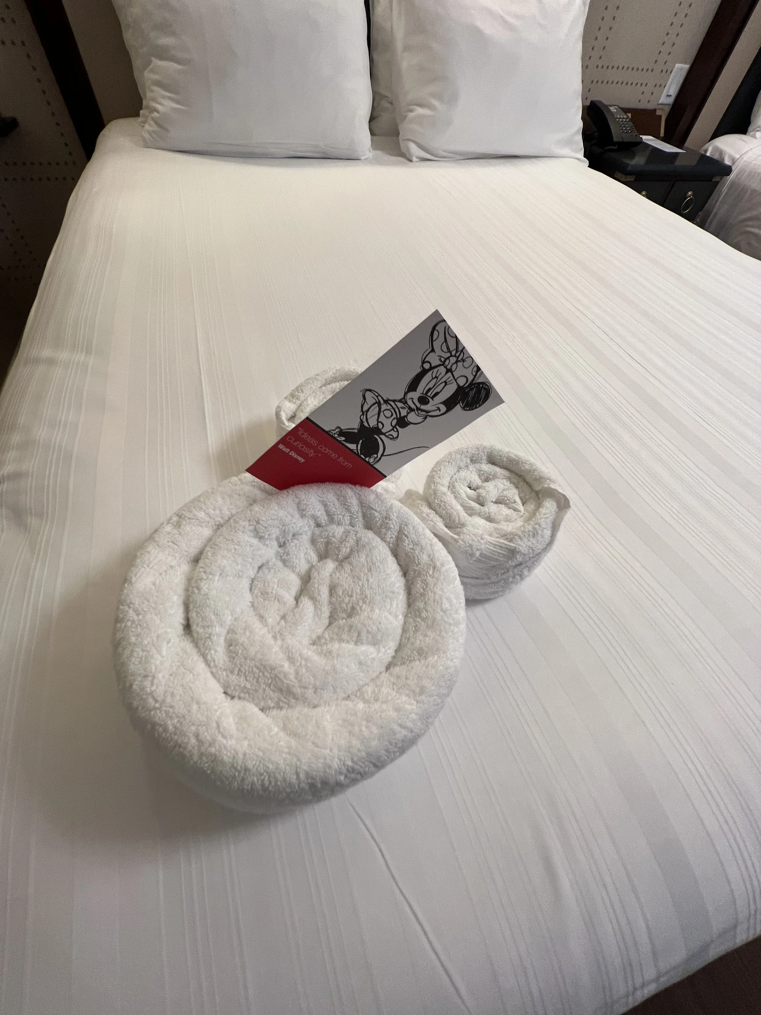 Hotel bed with two pillows and a towel folded into a rose shape, with a Mickey Mouse postcard inserted in the towel rose.