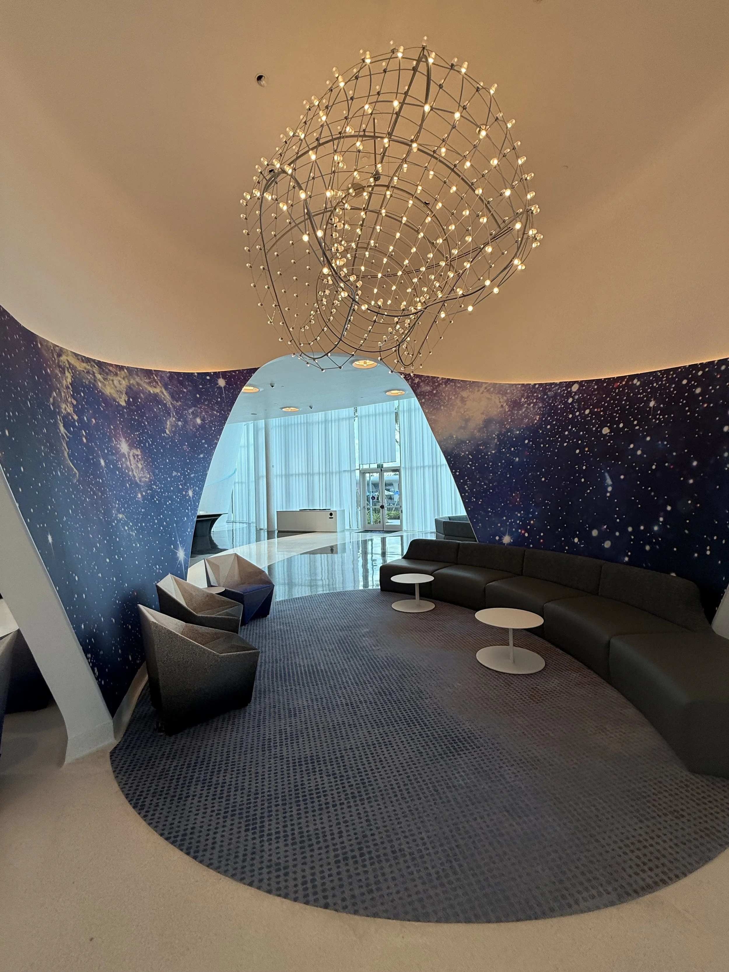 Modern hotel lounge with galaxy-themed wall mural, curved black sofa with small round white tables, and cluster of chairs near floor-to-ceiling windows, with a decorative chandelier hanging from the ceiling.