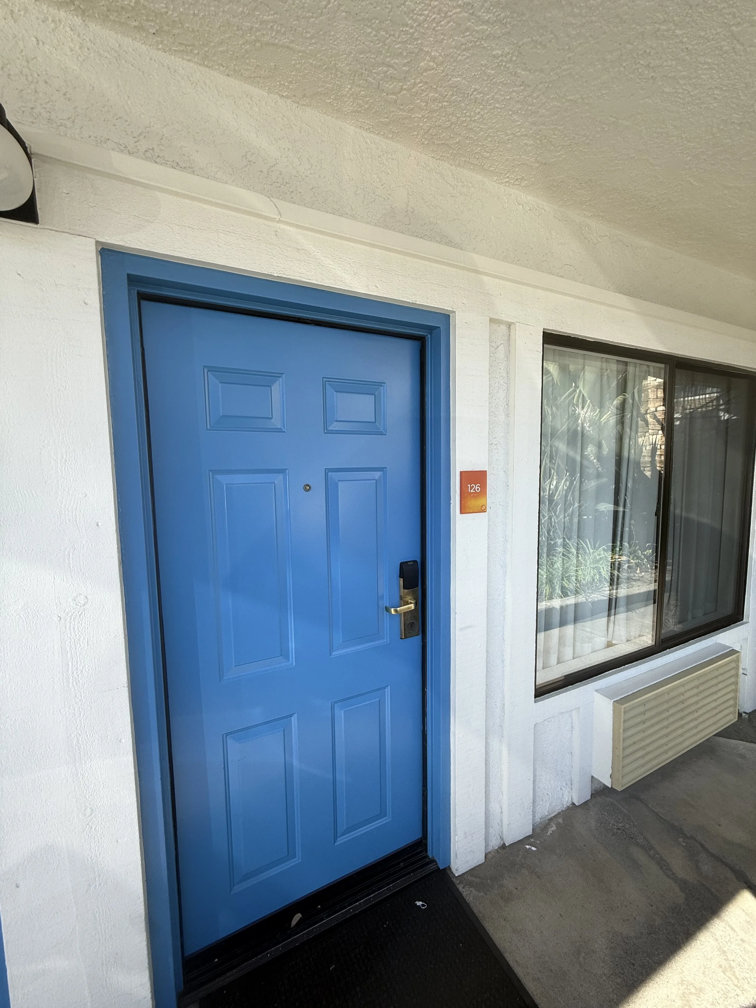An apartment door painted in bright blue with a black handle, set into a white wall, next to a window with reflective glass and beige curtain, a small orange number plaque reading 126, and an outdoor air conditioner below the window.