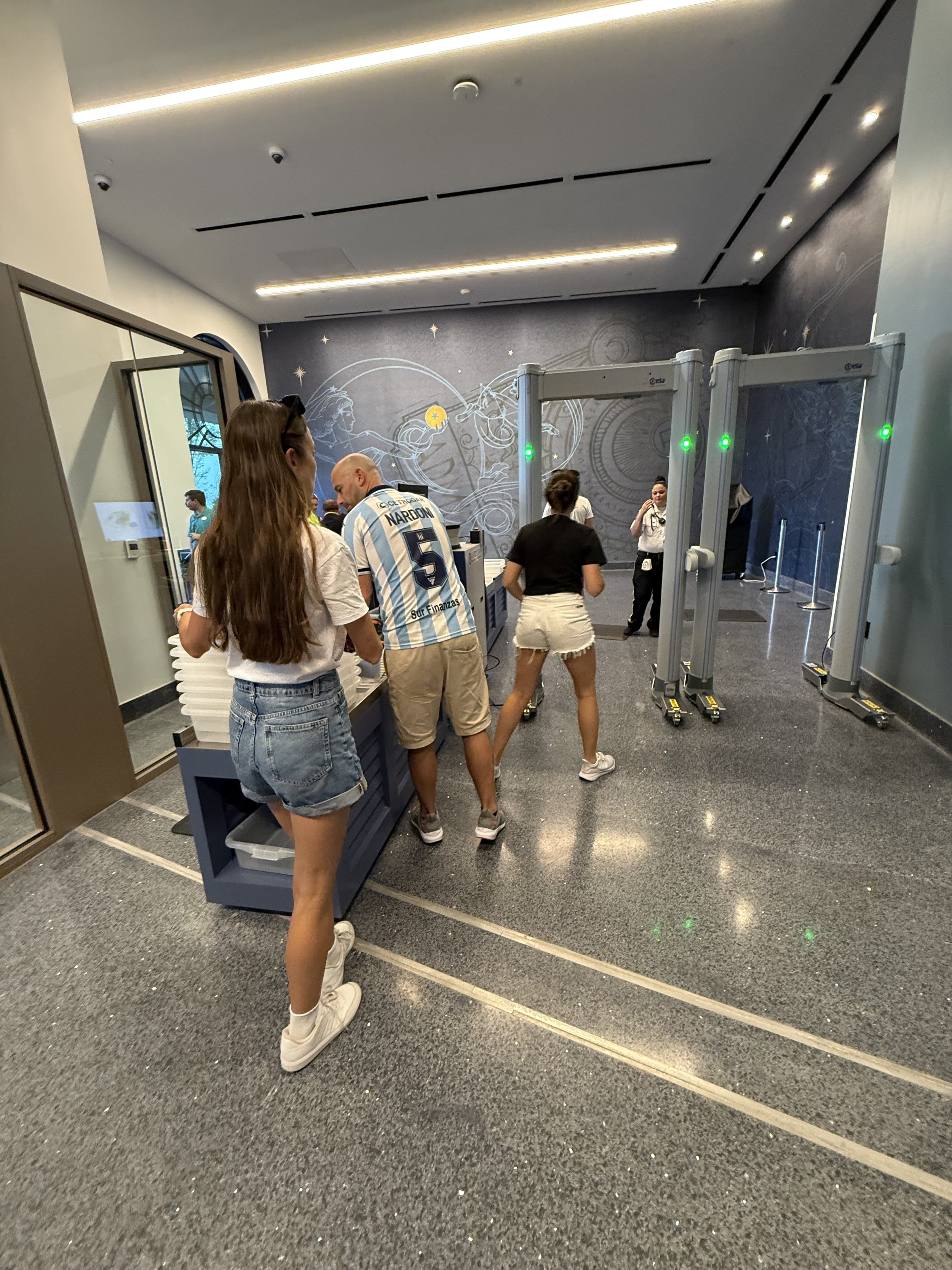 People in line at security checkpoint, scanning tickets or IDs at entrance to a facility with a space-themed wall art in the background.