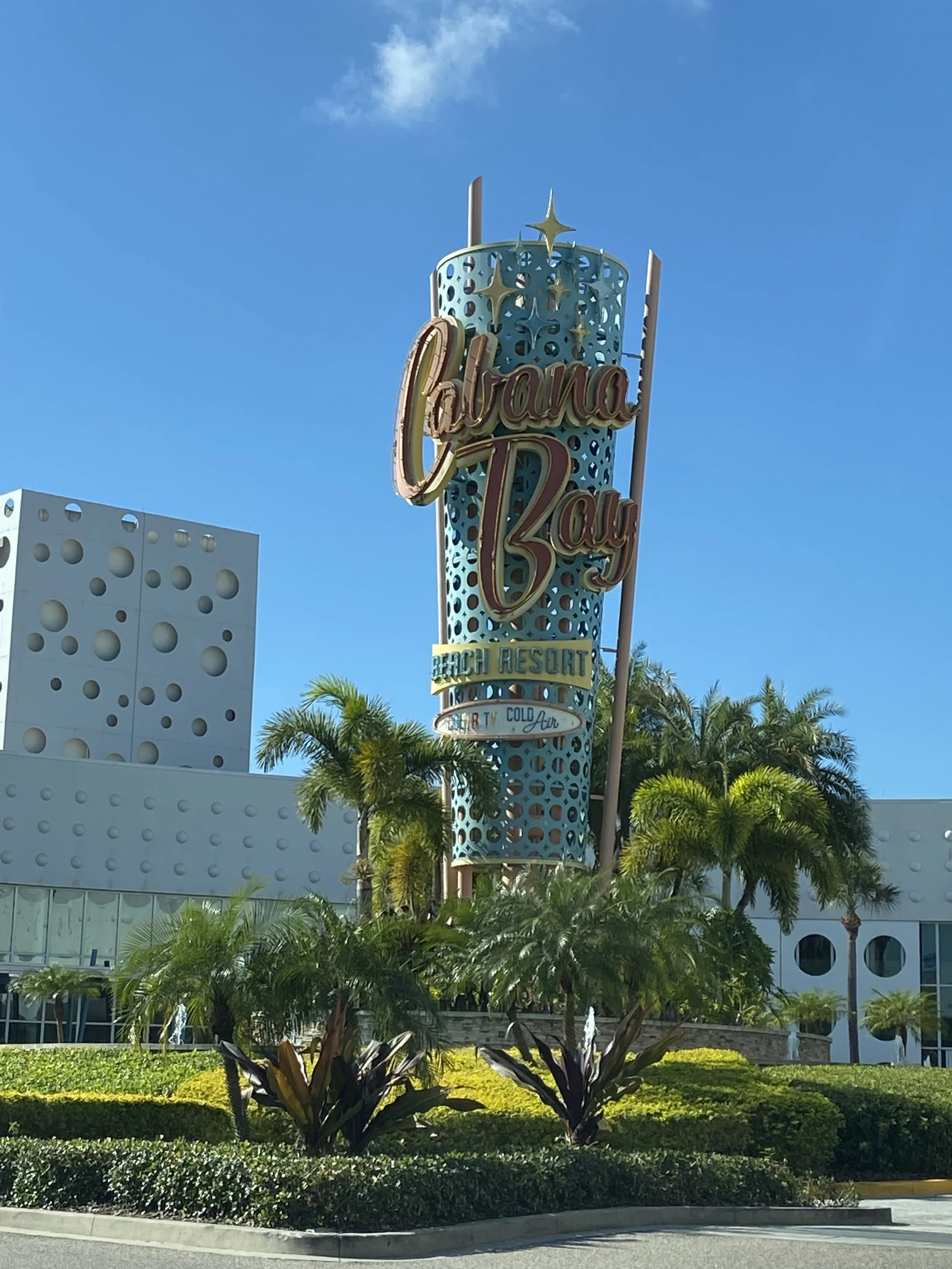 Sign for Catalina Bay, a beach resort with palm trees and modern white buildings in the background under a clear blue sky.