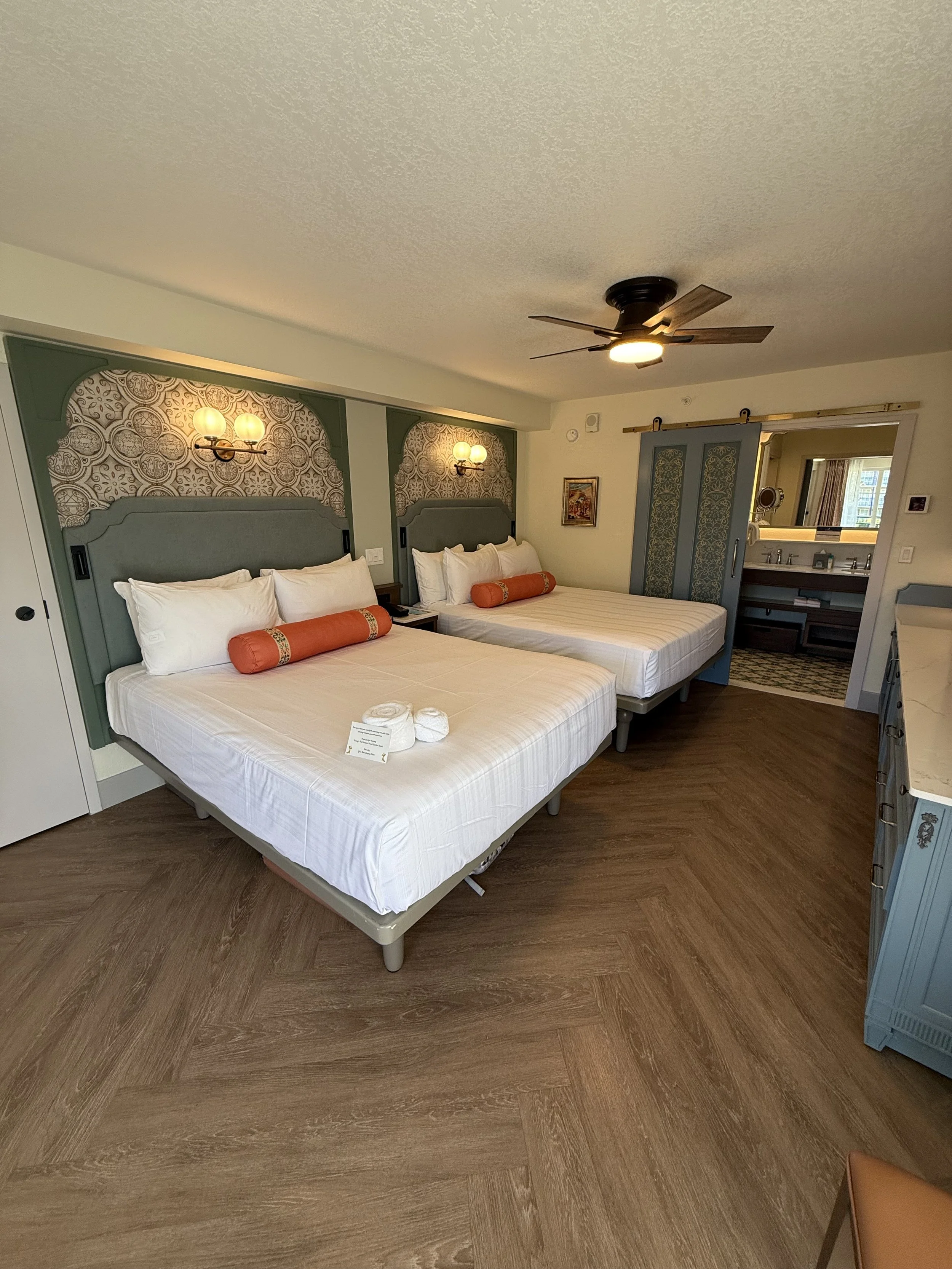 Two hotel beds with white linens and orange bolster pillows in a room with wood floor, green and patterned headboards, wall sconces, and a small bathroom in the background.
