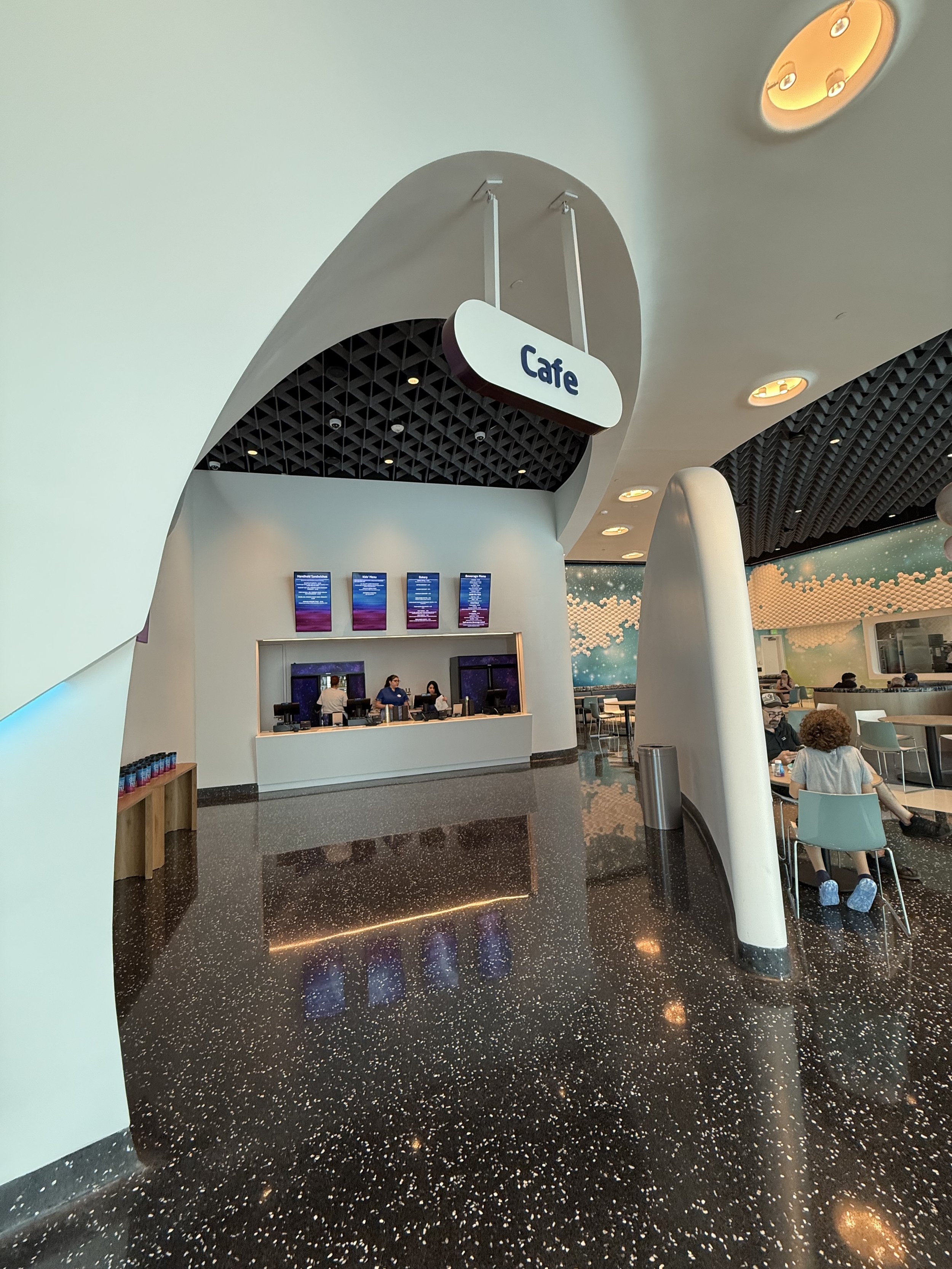 Interior of a modern cafe with a curved ceiling and a sign labeled 'Cafe'. A counter with staff, digital menu screens, and seating area with people dining.